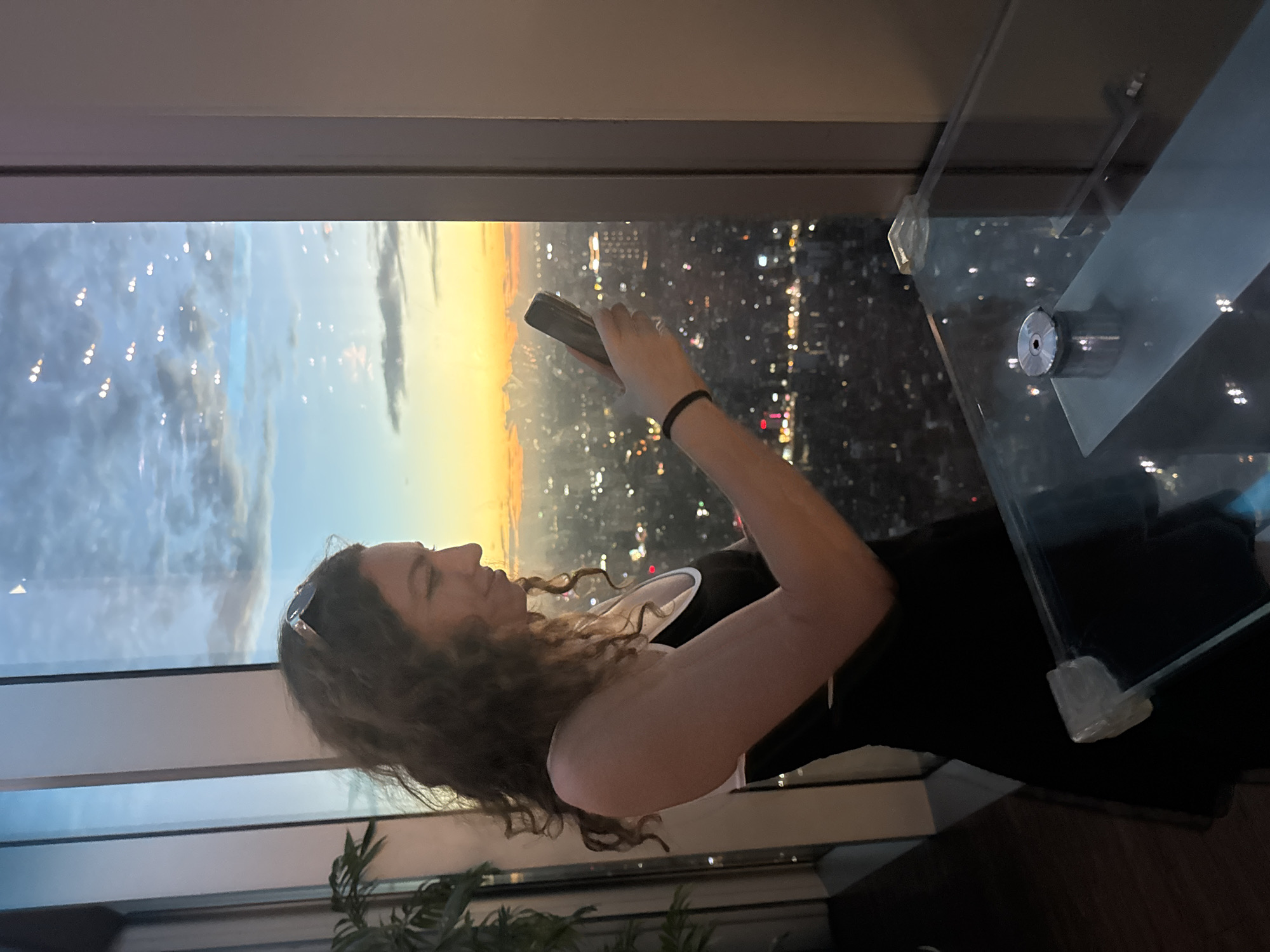 A woman with curly brown hair is standing next to a large window, looking at her phone. She is wearing a black dress with a white trim. The window offers a panoramic view of a city at sunset, with lights from buildings visible in the distance. The sky is a mix of orange, yellow, and blue, with some clouds. In the foreground, a glass table with a metal object on it is visible. The overall scene suggests an elevated viewpoint, possibly from a skyscraper, in a city like Hanoi, Vietnam, given the urban landscape and sunset.