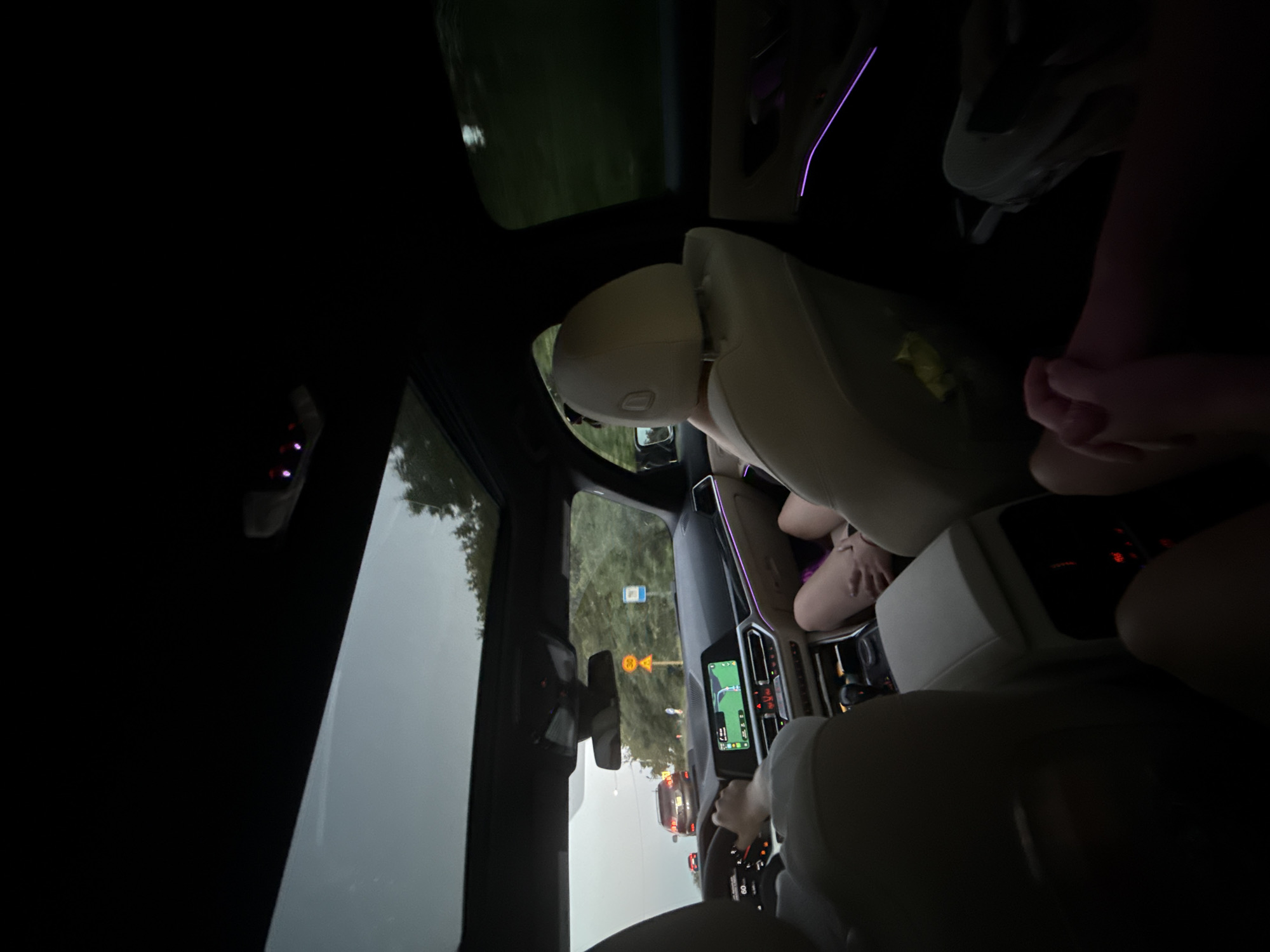 The image shows the interior of a car at dusk or night, with ambient lighting from the car's interior and exterior. The car is in motion, with other vehicles visible on the road ahead. The driver's hands are on the steering wheel, and a navigation screen is illuminated on the dashboard. Two passengers are visible in the back seat, with their legs extended. The car has a sunroof, which is open, and trees and foliage can be seen on either side of the road. There are traffic signs and streetlights visible on the road. The overall scene suggests travel during a transitional time of day. The location is identified as Fundul Galbenei, Moldova.