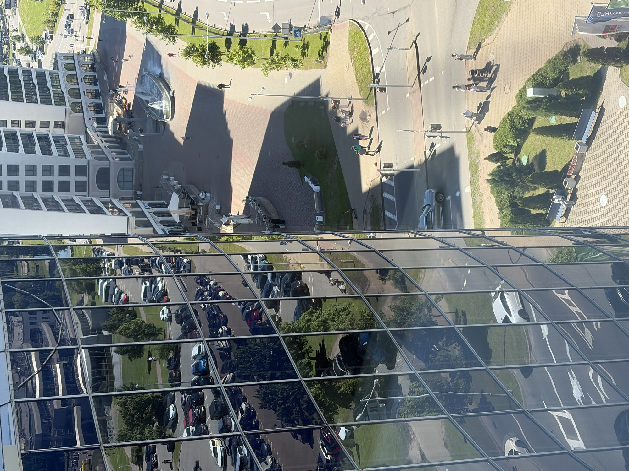 The image shows an urban scene in Barovlyany rural council, Belarus, viewed from a high angle looking down and slightly to the side. The foreground is dominated by the reflective glass facade of a modern building, which mirrors the surrounding environment. Reflected in the glass are rows of parked cars, trees, and parts of other buildings.
In the midground, a multi-story white building with arched windows is visible. In front of this building, there is a public space with a fountain in the center. People are walking in this area and along a paved walkway. To the right of this walkway, there is a road with several cars driving and parked. Green grassy areas and trees line the road.
Further into the background, more buildings and a road network are visible. The scene appears to be a well-developed urban or suburban area with modern architecture and infrastructure. The lighting suggests it is daytime, with strong sunlight casting shadows.