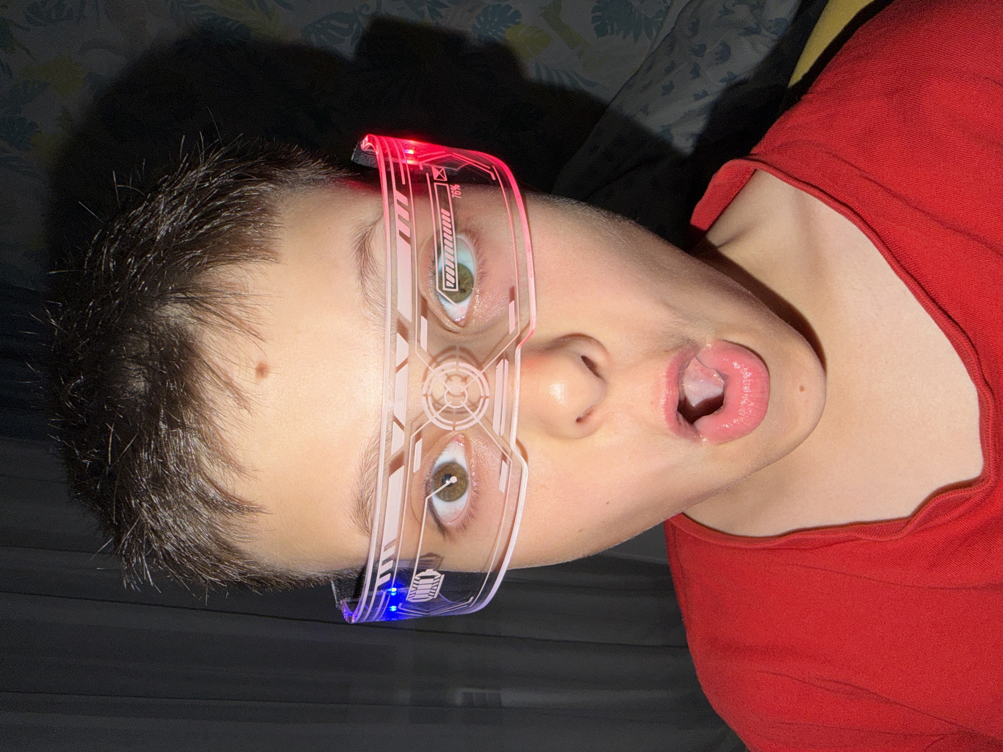 A young person, with dark hair and wearing a bright red shirt and futuristic LED glasses, looks directly at the camera with their mouth open in surprise. The glasses have a transparent frame with illuminated blue and red accents. The background consists of a dark curtain on the left and a patterned fabric on the right. The image appears to have been taken indoors.