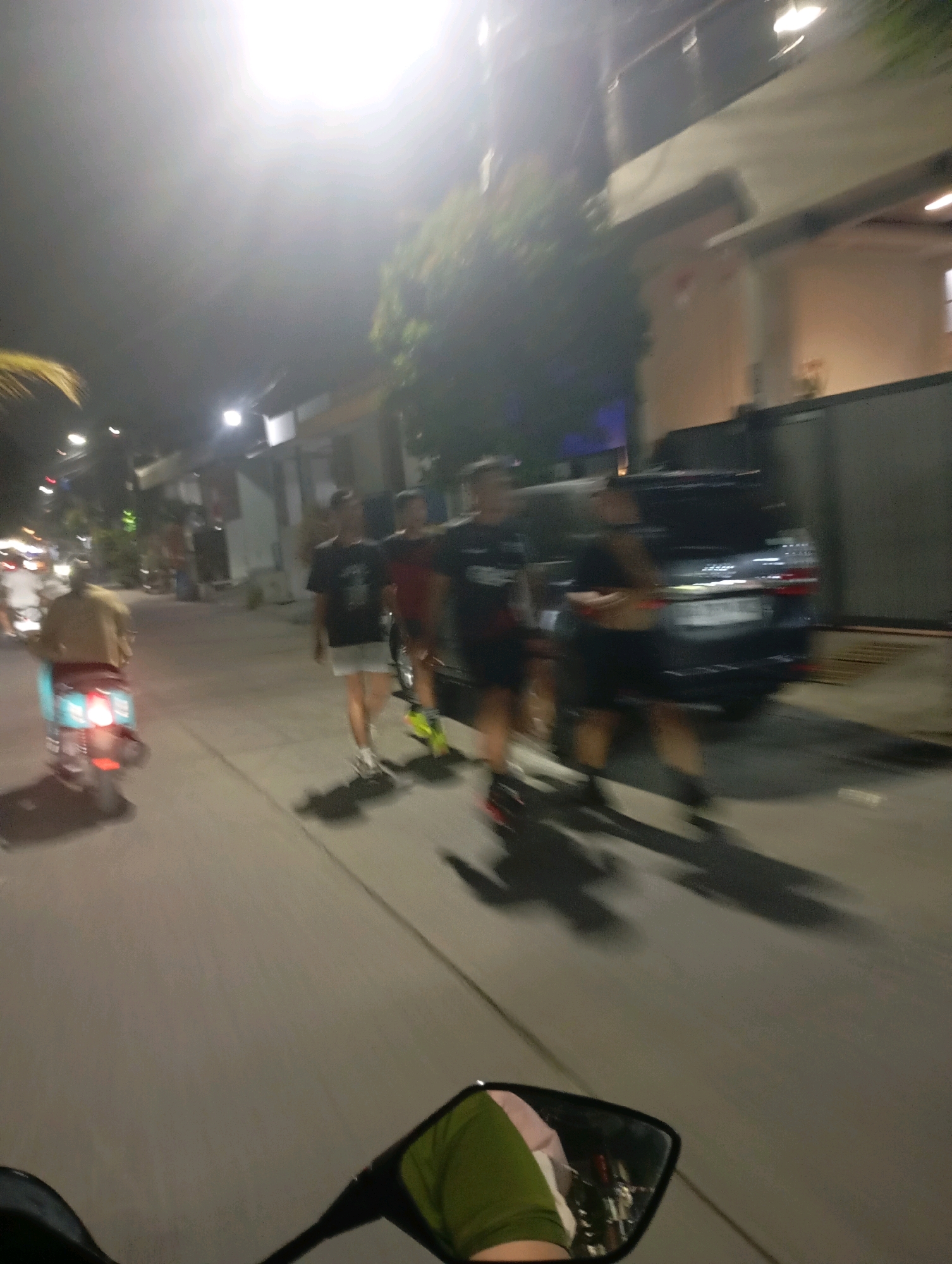 The image depicts a street scene at night in Babelan, Indonesia. In the foreground, the handlebar and a side mirror of a motorcycle are visible, indicating the perspective of someone riding or driving. The mirror reflects a person wearing a green shirt and possibly pink fabric. The background shows a street with several people walking. On the left, a person on a light blue scooter is heading away from the camera. Further down the street, a dark-colored SUV is parked. The environment is illuminated by streetlights and lights from buildings. Trees and foliage are also present along the street. The overall impression is of a typical evening street environment in an urban or suburban area.
