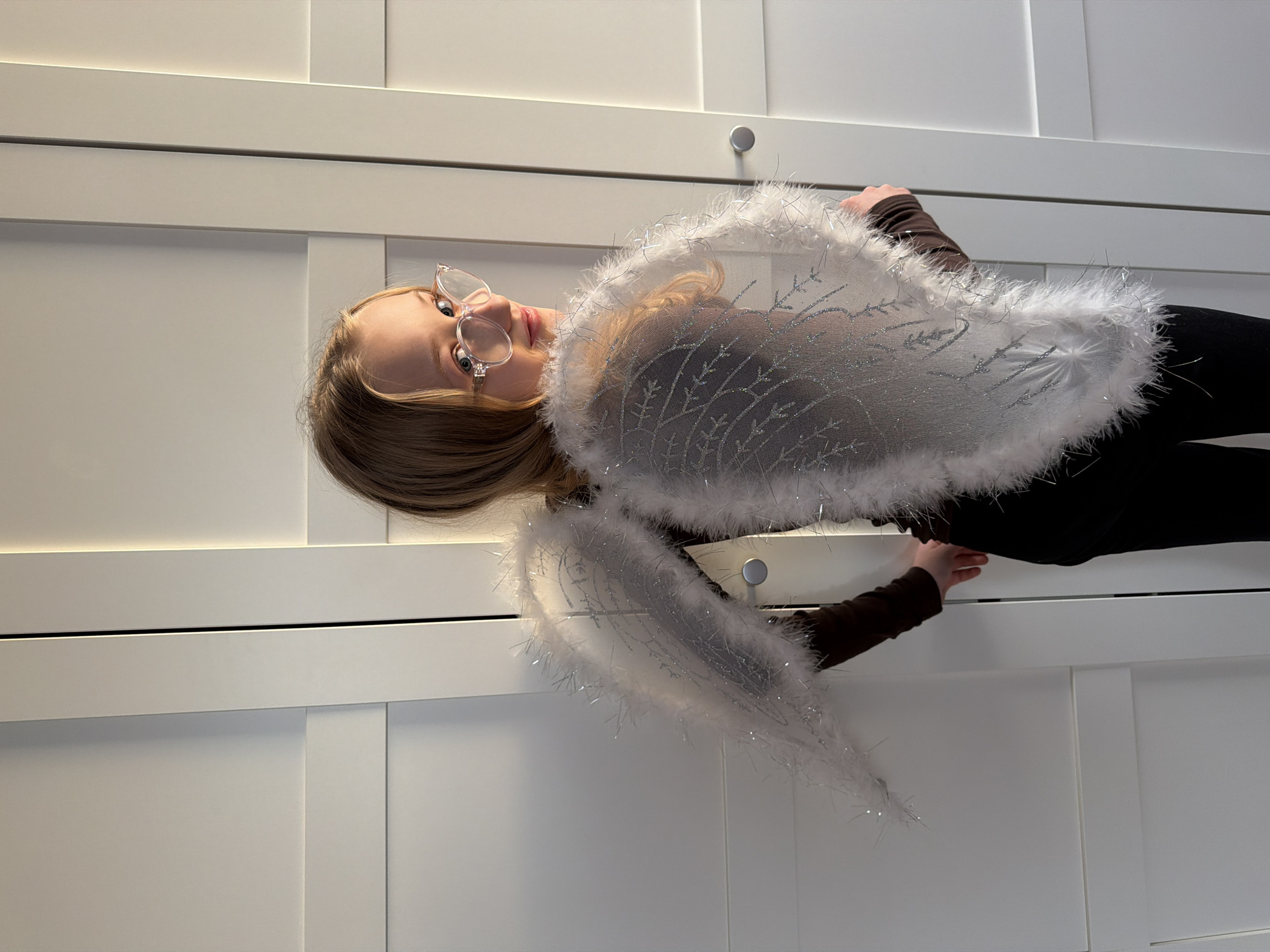 A young girl, wearing round eyeglasses and angel wings, poses in front of a white, paneled wall with silver circular door knobs. She is wearing a dark long-sleeved shirt and black pants. The location is likely indoors, but the specific location in Dreiliņi, Latvia is not discernible from the image.