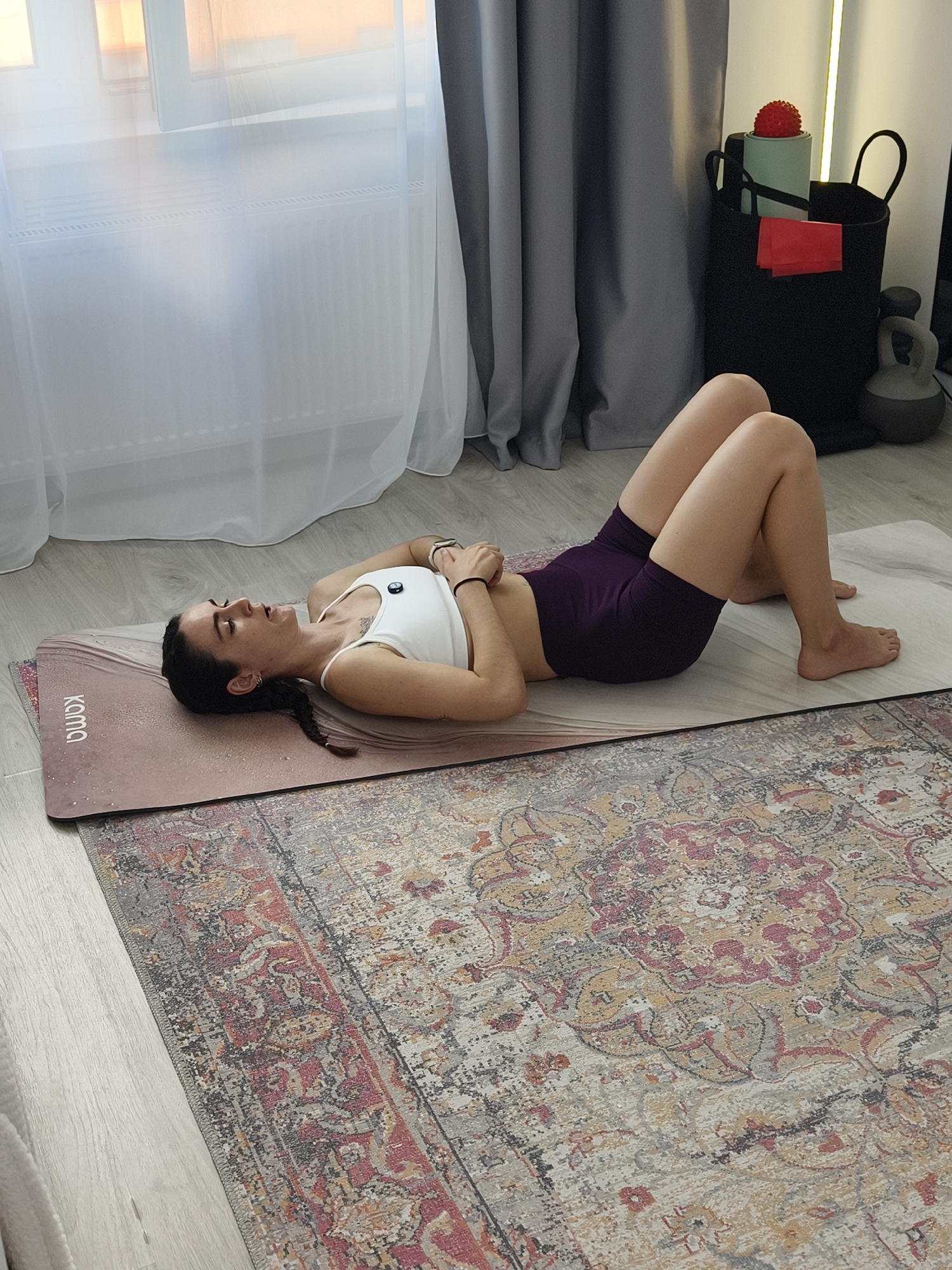 In Tyumen, Russia, a woman is shown lying on a yoga mat indoors. She is wearing a white sports bra and purple shorts. Her body is positioned on her back with her knees bent and feet flat on the mat. Her head is turned to the side, and her eyes are closed. The yoga mat is placed on a rug, which covers a portion of the wooden floor. In the background, a window with sheer curtains is visible, as well as a black storage bin containing various exercise equipment, including a red massage ball and resistance bands. A kettlebell and a lamp are also present in the background. The overall environment suggests a home fitness setting.