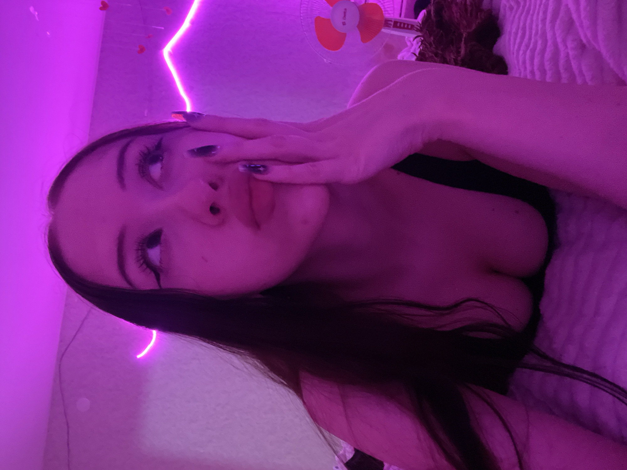 The image shows a young woman with long, dark hair, wearing a black top. She is lying down, propped up on her left arm, and her right hand is touching her face. She is looking upwards and to her left. The background is illuminated by pink neon lights, with a string of small red hearts hanging on the wall. A small fan is visible in the background to the right. The scene appears to be indoors, likely in a bedroom. No specific location within Votkinsk, Russia, can be determined from the image.