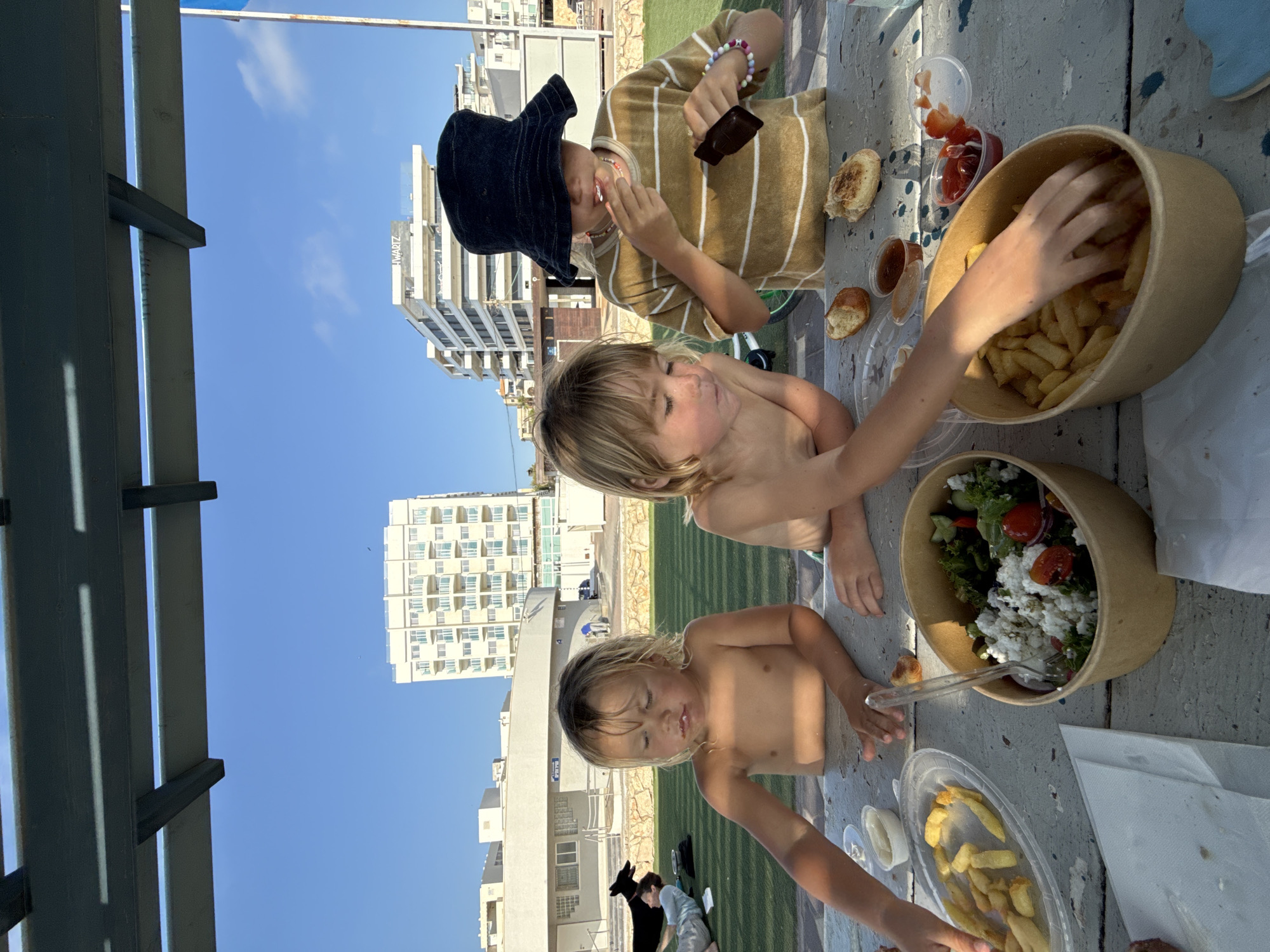 In Nahariya, Israel, three children are gathered around a picnic table, enjoying an outdoor meal. The table is laden with food, including bowls of fries, a salad with feta cheese and tomatoes, and what appears to be bread rolls. One child, wearing a striped t-shirt and a dark bucket hat, is actively eating. Another child, shirtless, is reaching for fries from a large container. A third child, also shirtless, is seated in the foreground, looking down at the table. In the background, a black dog lies on the grass next to a person. Further in the distance, two large, multi-story buildings with balconies are visible against a clear blue sky. The scene is set under a wooden pergola, providing shade.