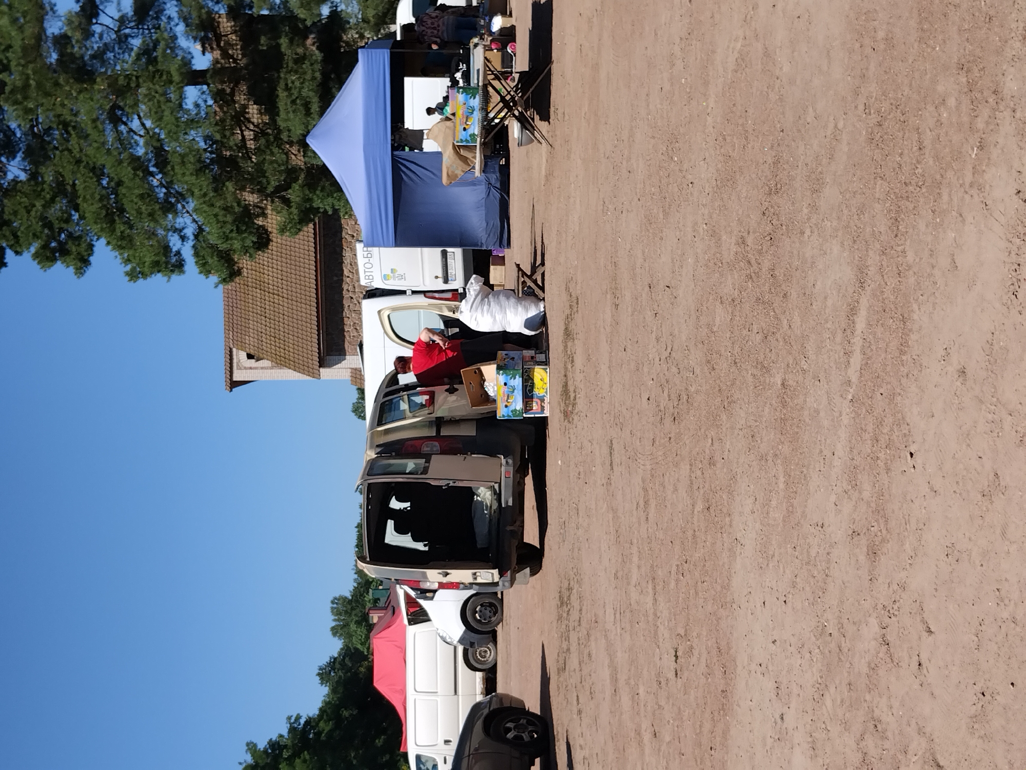 The image captures an outdoor market scene on a sunny day, likely in Irshansk, Ukraine, given the context. The ground is a dry, dirt lot with tire tracks visible. In the mid-ground, a beige van with its rear doors open is parked. A person in a red shirt and black shorts is standing beside the open van, and several cardboard boxes are placed on the ground in front of it. To the right of the van, a large white bag is positioned next to a small wooden stool.
Further to the right, a blue pop-up tent is set up. Beneath the tent, a foldable table displays several items, including boxes of what appear to be bananas. Two people are visible near this table, one standing and the other seated. In the background, more vans are parked, one white with a red top, and another white van with text that includes "АВТО-БР". A building with a tiled roof and stone elements is visible behind the tents and vans. Lush green trees and a clear blue sky complete the environment.