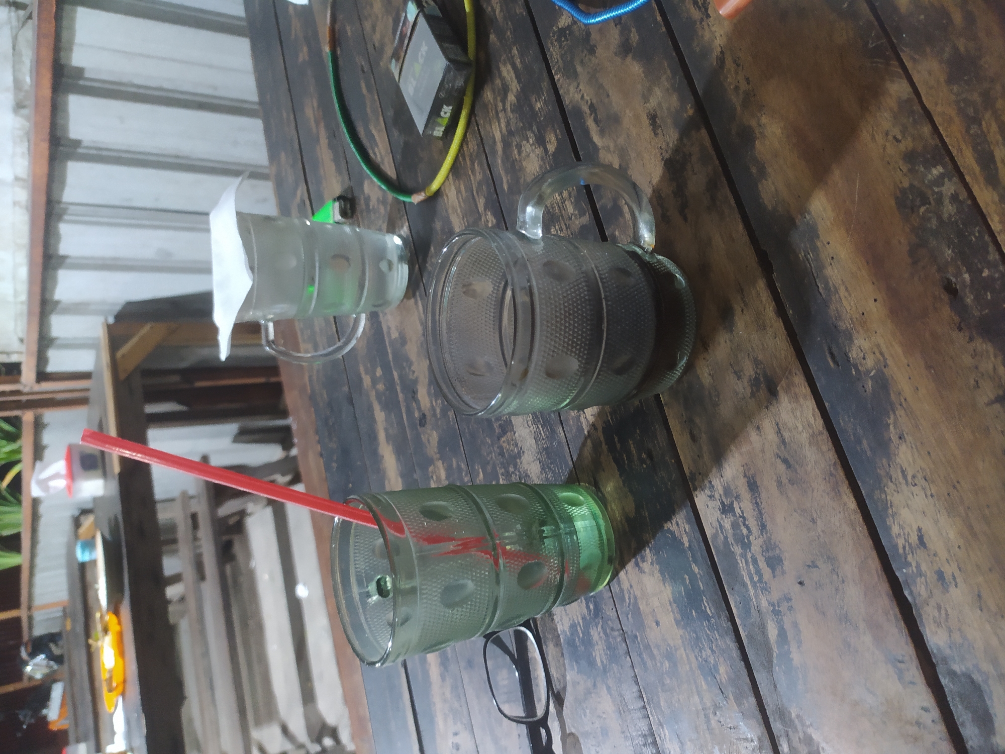 An outdoor cafe table in Tuntang, Indonesia, is set with two drinks, a cigarette pack, a lighter, and a pair of glasses. A green glass with a red straw contains a clear liquid. To its right, a clear mug holds a dark liquid, likely coffee. A pack of "BLACK" cigarettes and a green and yellow hose are visible, suggesting a shisha pipe. The table is made of rough, dark wood, with shadows suggesting it is nighttime. In the background, more tables and chairs are visible, along with the corrugated metal wall of a building. The scene suggests a casual evening gathering.