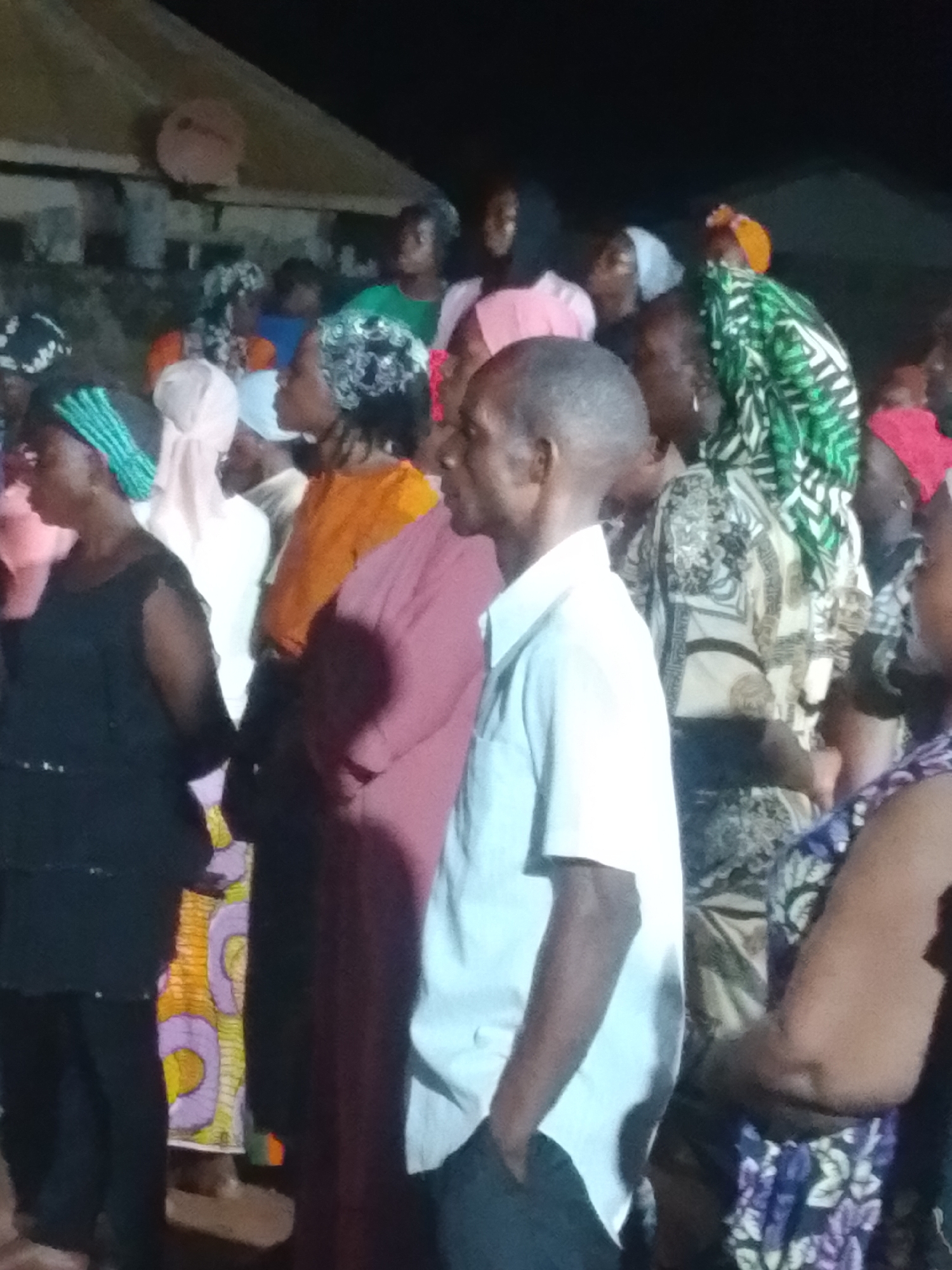 An outdoor gathering is depicted at night, with a group of people, predominantly women wearing headscarves and traditional African clothing, assembled. The atmosphere appears to be one of community or a social event. The lighting is dim, suggesting it's nighttime, and the background is indistinct, showing glimpses of a structure that might be a building or shelter. In the foreground, a man in a light-colored shirt stands in profile, facing right. To his left, women are visible in various colorful outfits, including a woman in a black dress and a teal headscarf, and another in an orange garment with a patterned headscarf. The overall scene is densely populated with individuals, and the focus is on the gathering itself.