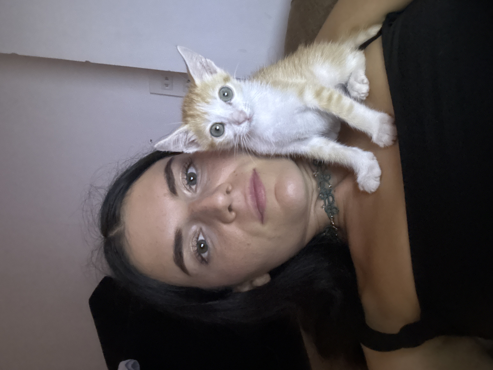 A woman lies on her back with an orange and white kitten resting on her head and upper chest. The woman has dark hair, light skin, and light green eyes. She is wearing a dark-colored top and a beaded necklace. The kitten is small and appears to be looking at the camera. The background is a plain wall with an electrical outlet visible. The location is not discernible from the image.