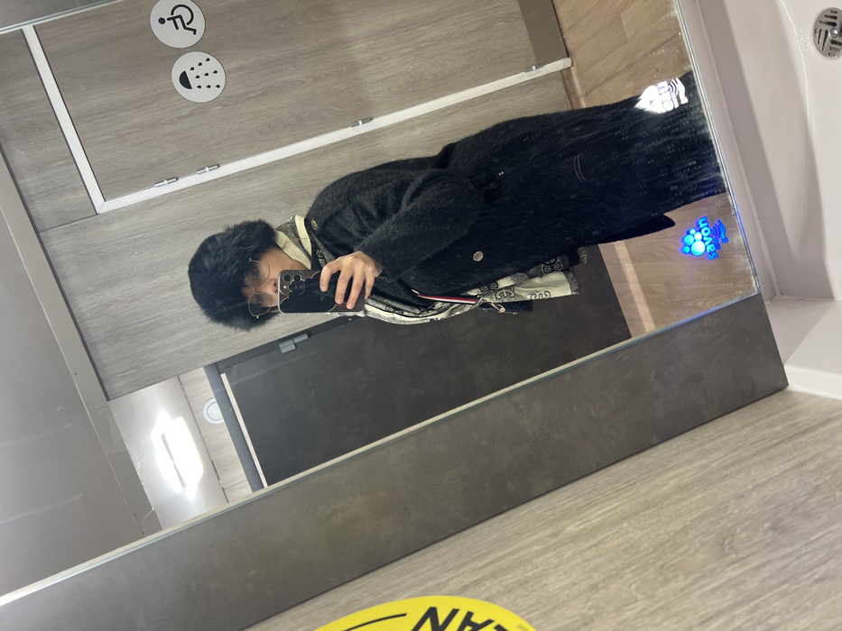 A person in a black furry hat and round glasses takes a mirror selfie. They wear a dark, textured coat over a lighter-colored shirt and a patterned scarf. The bathroom setting includes gray walls, a white sink, and signage indicating facilities for disabled people. A "clean" sign is visible on the floor, and a hand soap dispenser is integrated into the mirror.
