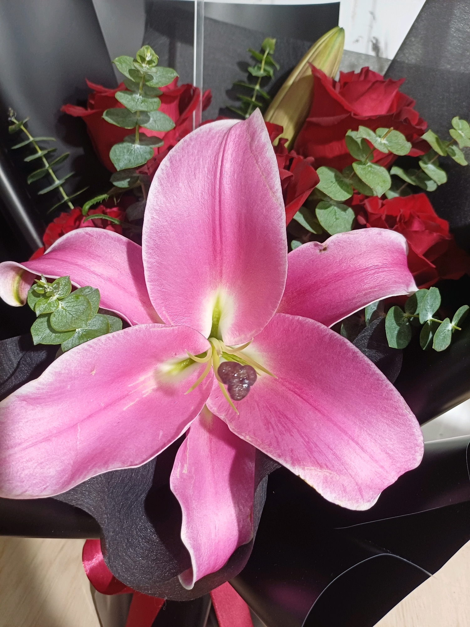 A close-up, eye-level shot showcases a bouquet of flowers, predominantly featuring a large, vibrant pink lily in the foreground. The lily's petals are wide and soft, with delicate pink hues that lighten towards the edges. At the center of the lily, visible stamens are clustered, with a dark, almost black pollen structure at their tips. Behind the lily, several deep red roses are interspersed with sprigs of green eucalyptus leaves. The bouquet is wrapped in black paper, with a red ribbon tied at the bottom, suggesting it is a gift. The background is out of focus, hinting at a neutral, possibly indoor setting with a light-colored surface below and a textured, reflective surface behind. There are no people or specific city landmarks identifiable within the image. The lighting appears to be natural, highlighting the textures and colors of the flowers.