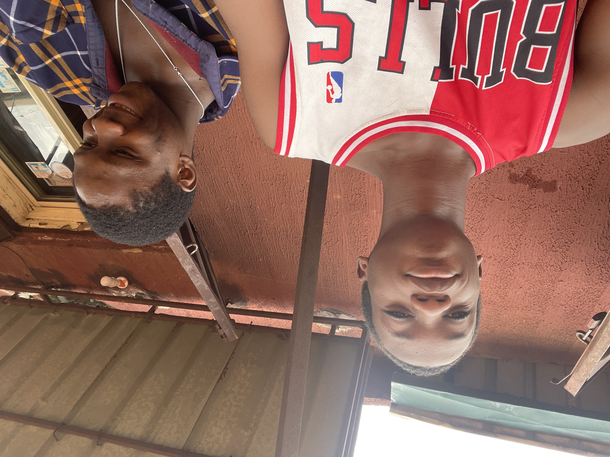 Two young males are pictured outdoors, positioned in the foreground. The male on the left, facing forward, wears a red and white Chicago Bulls basketball jersey with the word "BULLS" prominently displayed. He has short black hair and a neutral expression. To the right, the second male is turned away, looking downwards and smiling. He is wearing a blue, yellow, and white plaid shirt with a maroon collar and a silver chain. The background reveals a reddish-brown textured wall, a corrugated metal roof supported by metal beams, and a glimpse of a window with some signage on the right. The overall scene suggests an informal outdoor setting, potentially in a residential or commercial area.