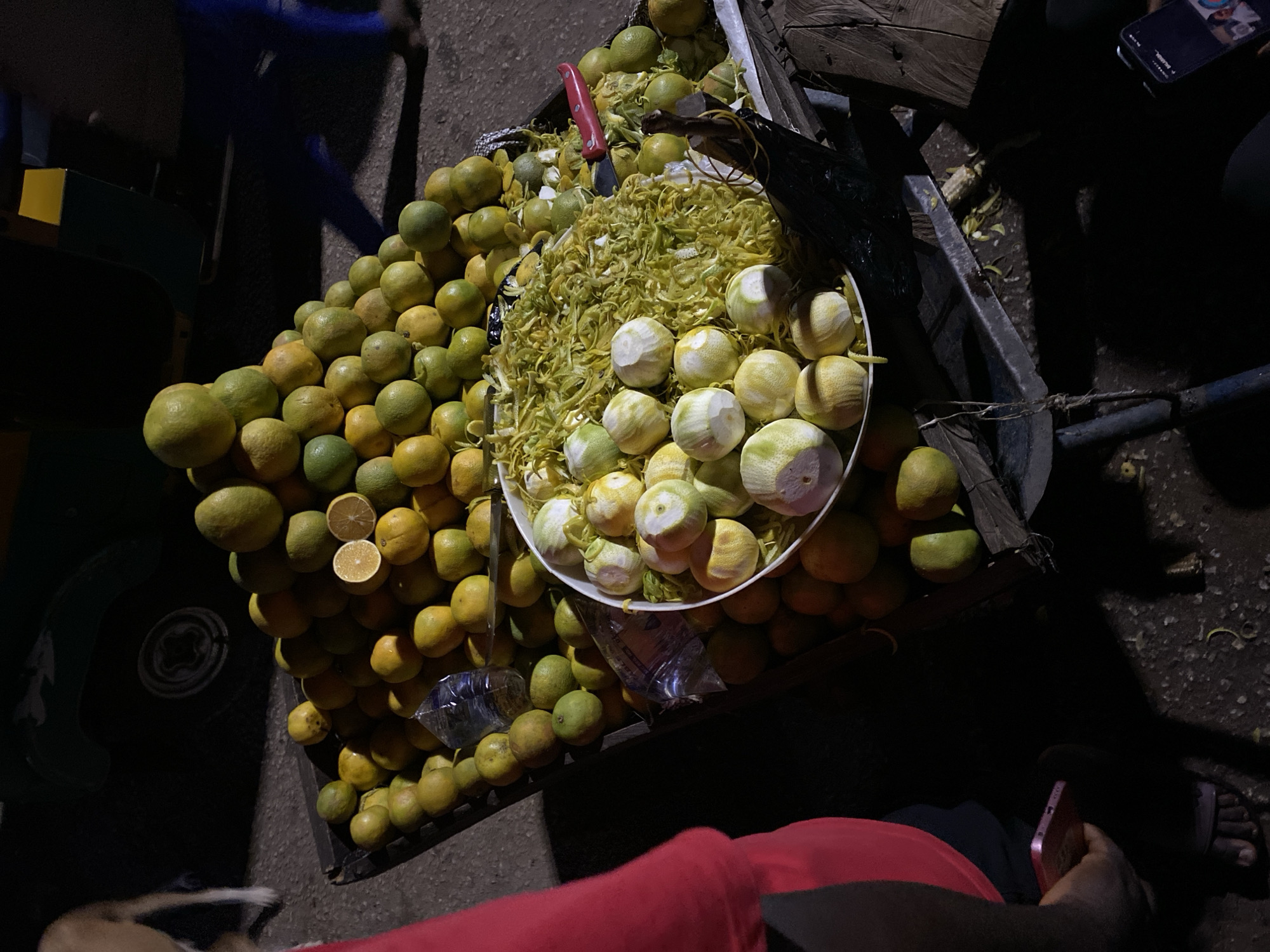 The image depicts a fruit stand in Bukuru, Nigeria, at night. The main focus is on a large pile of green and yellow citrus fruits, some whole and some partially peeled, with the peels collected into shredded piles. A red-handled knife is visible among the fruit. The fruit is arranged on a cart, part of which is obscured by darkness. A person in red clothing and sandals is holding a mobile phone in the lower right corner. Another person's legs are visible on the left of the cart.