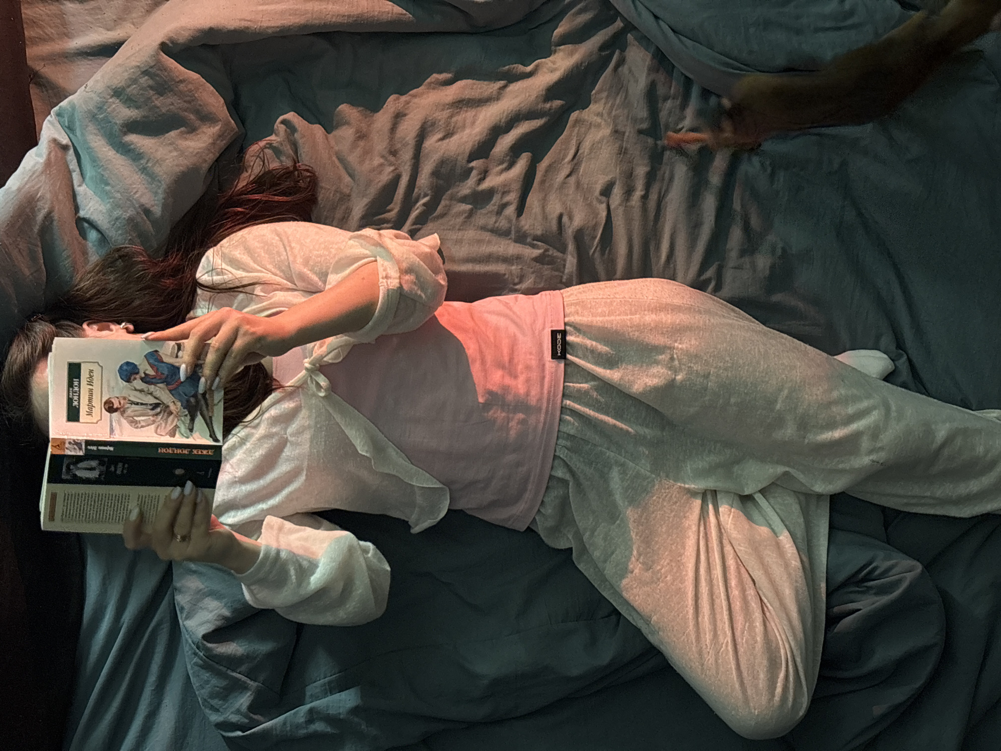 A woman lies on a rumpled, grey-blue bed covered in shadows. She is wearing a light-colored, loose-fitting outfit with a pink top underneath. She is holding a book titled "Юидон Мартин Иден Джек Лондон" ("Юидон" is a Russian transliteration of "Юидон" and likely a misspelling for "Иден" (Eden)). The title is partially obscured. The woman's face is hidden behind the book. A hand (or arm) is visible in the upper right corner of the frame, extending onto the bed. The photo seems to have been taken in dim lighting. The location is Chaikovsky, Russia.