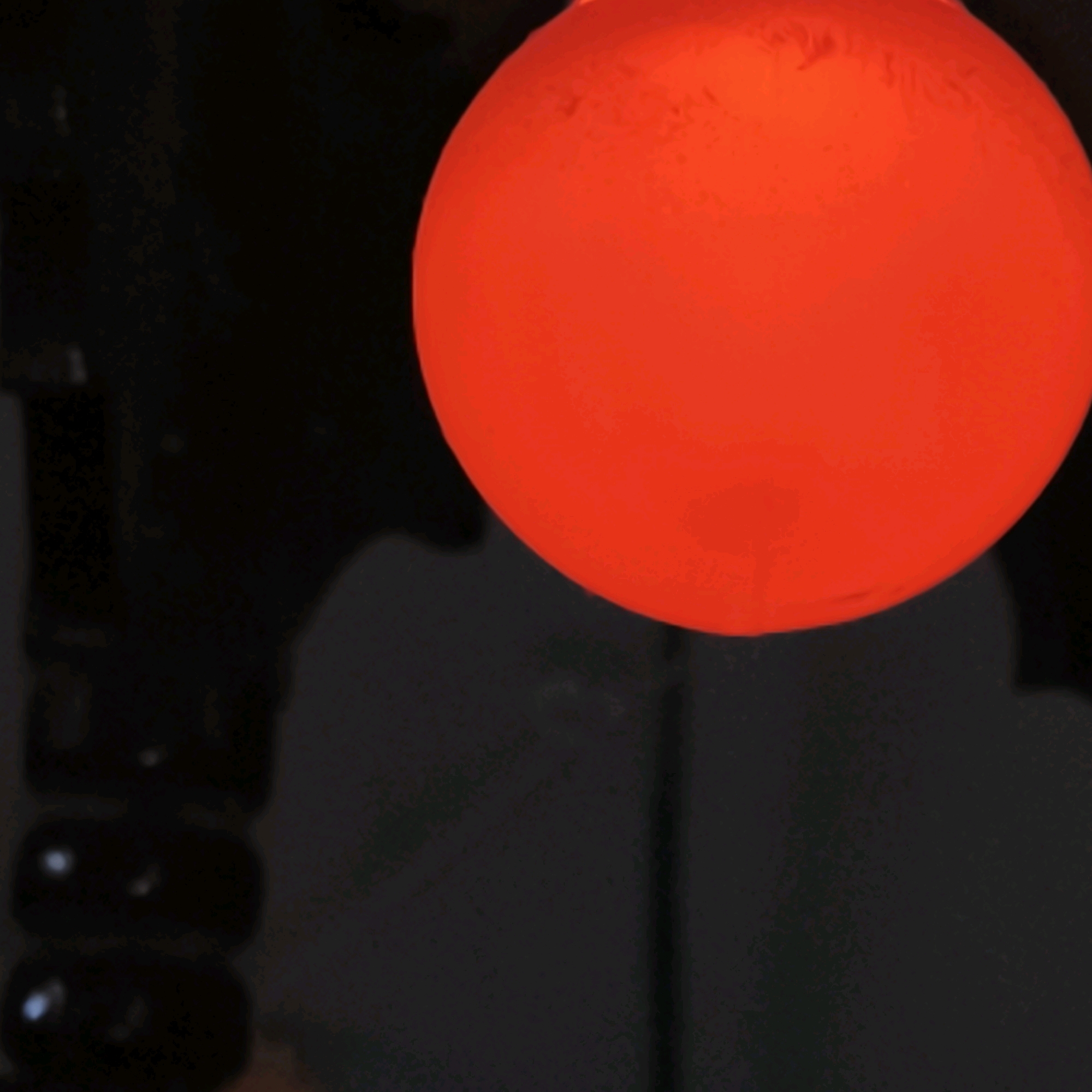 The image is a close-up shot of a glowing red orb against a dark background. The orb appears to be illuminated from within, with subtle variations in brightness and texture on its surface. The dark background is indistinct and appears to have some vertical, elongated shapes on the left side, possibly suggesting a dark, textured surface or objects. The lighting is dramatic, highlighting the vibrant red of the orb and creating deep shadows. There is no discernible location in Mumbai, India, or any people or actions depicted.