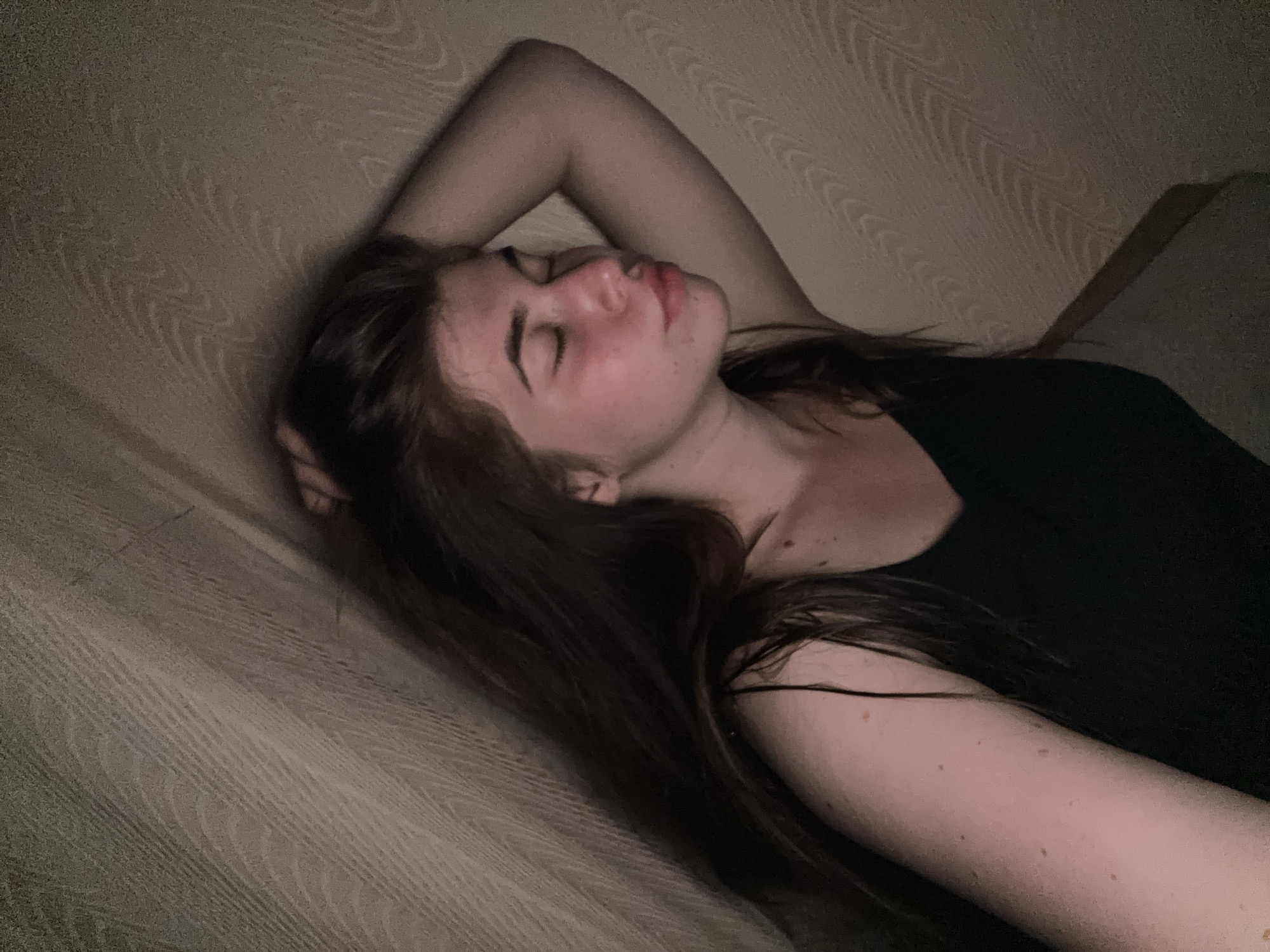 A young woman with long, dark brown hair is lying down with her eyes closed and her right arm raised behind her head. She is wearing a black tank top. Her skin is fair with some freckles visible on her arm. The background appears to be a wall with a textured pattern. The scene is dimly lit, creating shadows that obscure some details. The woman's facial expression suggests she is relaxed or sleeping. There are no other people or objects visible in the frame. The location is in the city of Мещёры, Russia.