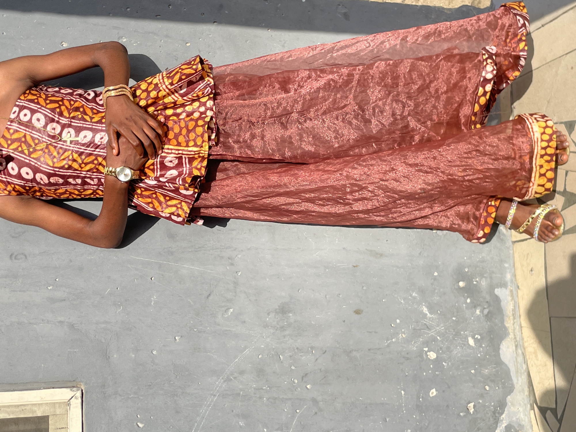 An overhead shot shows a person lying or reclining outdoors on a grey, textured concrete surface, likely in Badagry, Nigeria. The individual, with dark skin, is dressed in a vibrant patterned top or bodice featuring brown, orange, and white circular and leaf-like designs. This is paired with a flowing, translucent reddish-brown skirt or wrapper, which appears crinkled and reflects light.
The person's hands are clasped together over their midsection. They wear a gold-colored watch with a round face and multiple gold-colored bangles on their left wrist. A delicate gold-colored necklace with a small cross pendant is visible on their chest. Gold-colored sandals with white beads are worn on their right foot, which is partially visible. The hem of the skirt also features a border matching the pattern of the top.
The scene is set outdoors under bright, direct sunlight, evidenced by the sharp shadows cast by the person and their attire onto the ground. The grey concrete ground shows signs of wear, with visible scratches, varying tones, and small chipped areas. In the bottom right corner, a section of light-colored, possibly cream or beige, tiled flooring is visible next to the concrete. A small white rectangular object, potentially a window frame, is visible in the bottom left corner. The clear, sunny weather suggests daytime. No text is visible in the image.