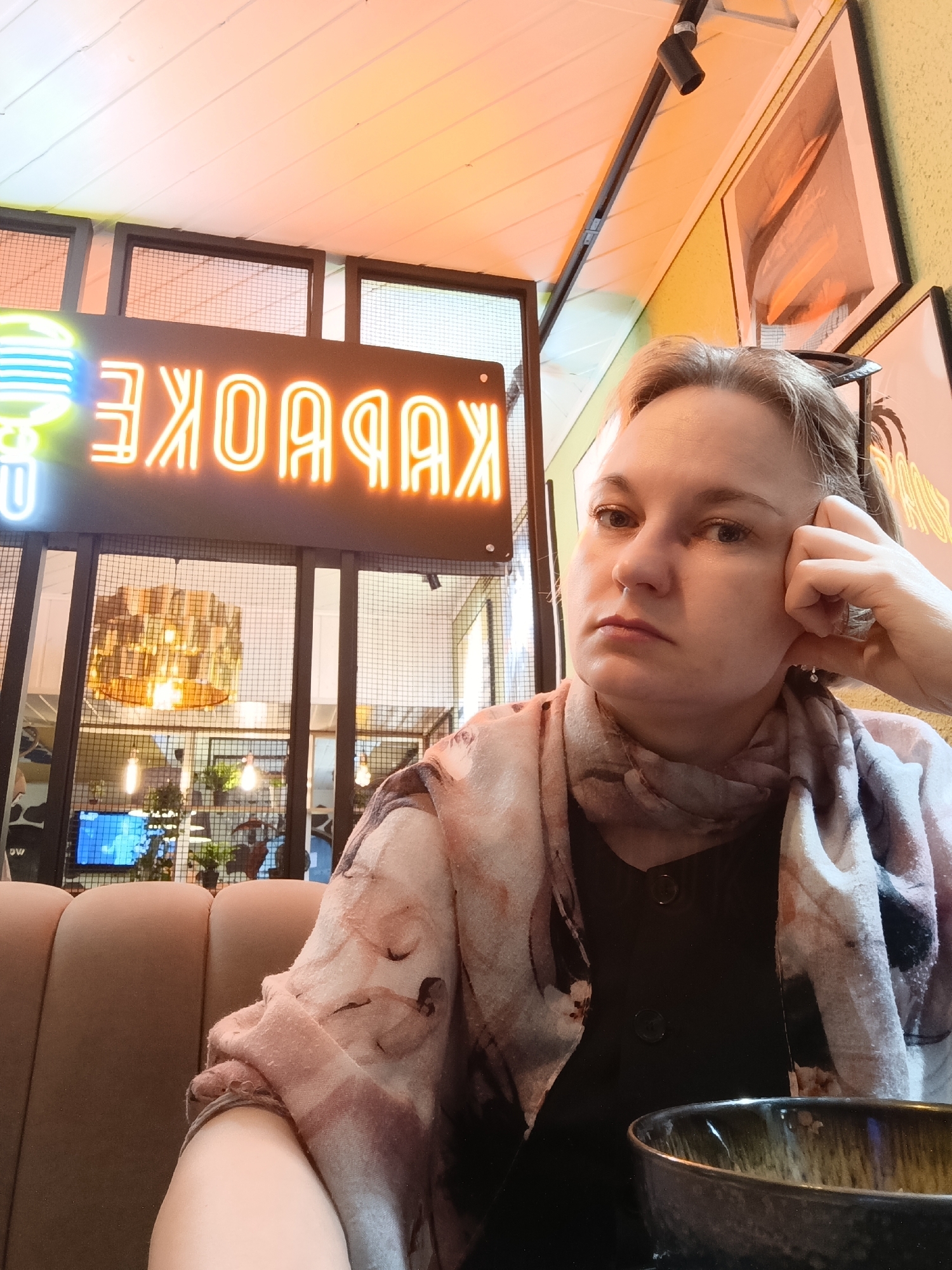 The image features a woman seated indoors at what appears to be a cafe or restaurant in Chaikovsky, Russia. She has fair skin, light-colored hair, and wears a patterned scarf in muted pinks and browns over a dark top. She is positioned in the foreground, leaning her head on her right hand, which displays a ring. Her expression is neutral to slightly pensive as she looks towards the camera.
In the immediate foreground, a dark bowl with a glazed finish rests on the table. The background reveals the interior setting with brown upholstered booth seating. A prominent neon sign, brightly lit in orange, is visible to the left and spells "KARAOKE" in reverse due to the likely selfie perspective. Behind the sign, large windows with a metal grid pattern separate areas, through which glimpses of other sections, including warm ambient lighting and a television screen displaying blue hues, can be seen. The overall lighting within the space is warm, with an orange tint, particularly from the neon sign. The scene suggests a relaxed atmosphere, possibly during the evening.