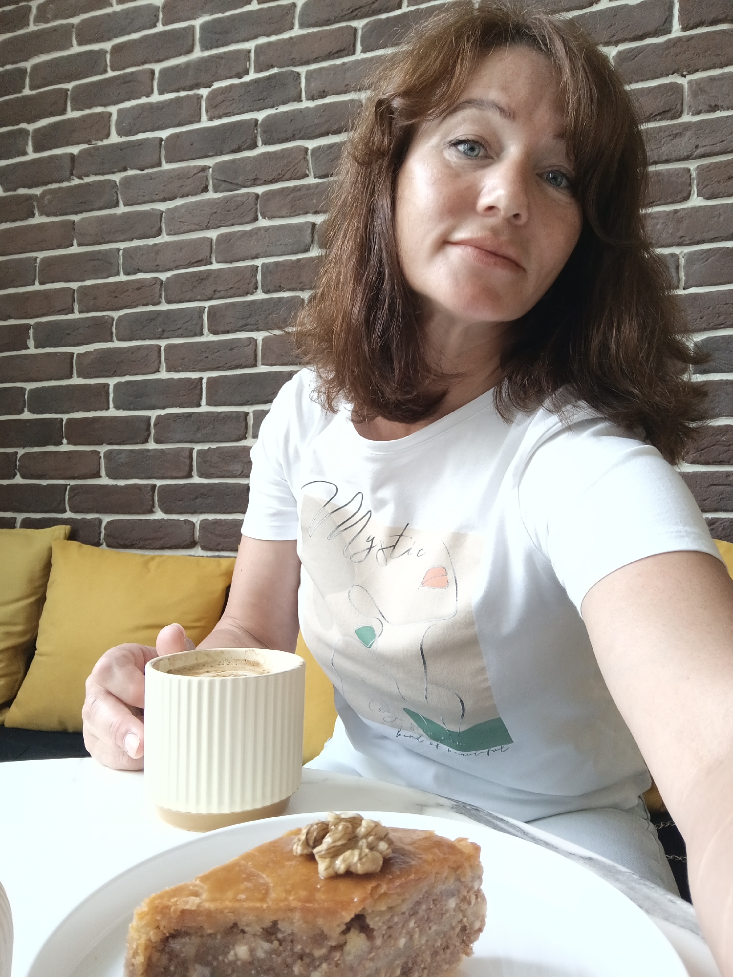 A medium-shot, eye-level, selfie shows a woman with auburn hair, light blue eyes, and fair skin sitting at a white table in front of a dark brown brick wall. She is wearing a white t-shirt with a graphic print of a face and text that reads "kind of beautiful." She is holding a light beige ceramic mug filled with coffee in her right hand. In the foreground, on a white plate, is a piece of baklava topped with a walnut. Yellow pillows are visible on a couch behind her to the left. The scene appears to be in an indoor cafe or dining area. The provided context states the location is Sochi, Russia.