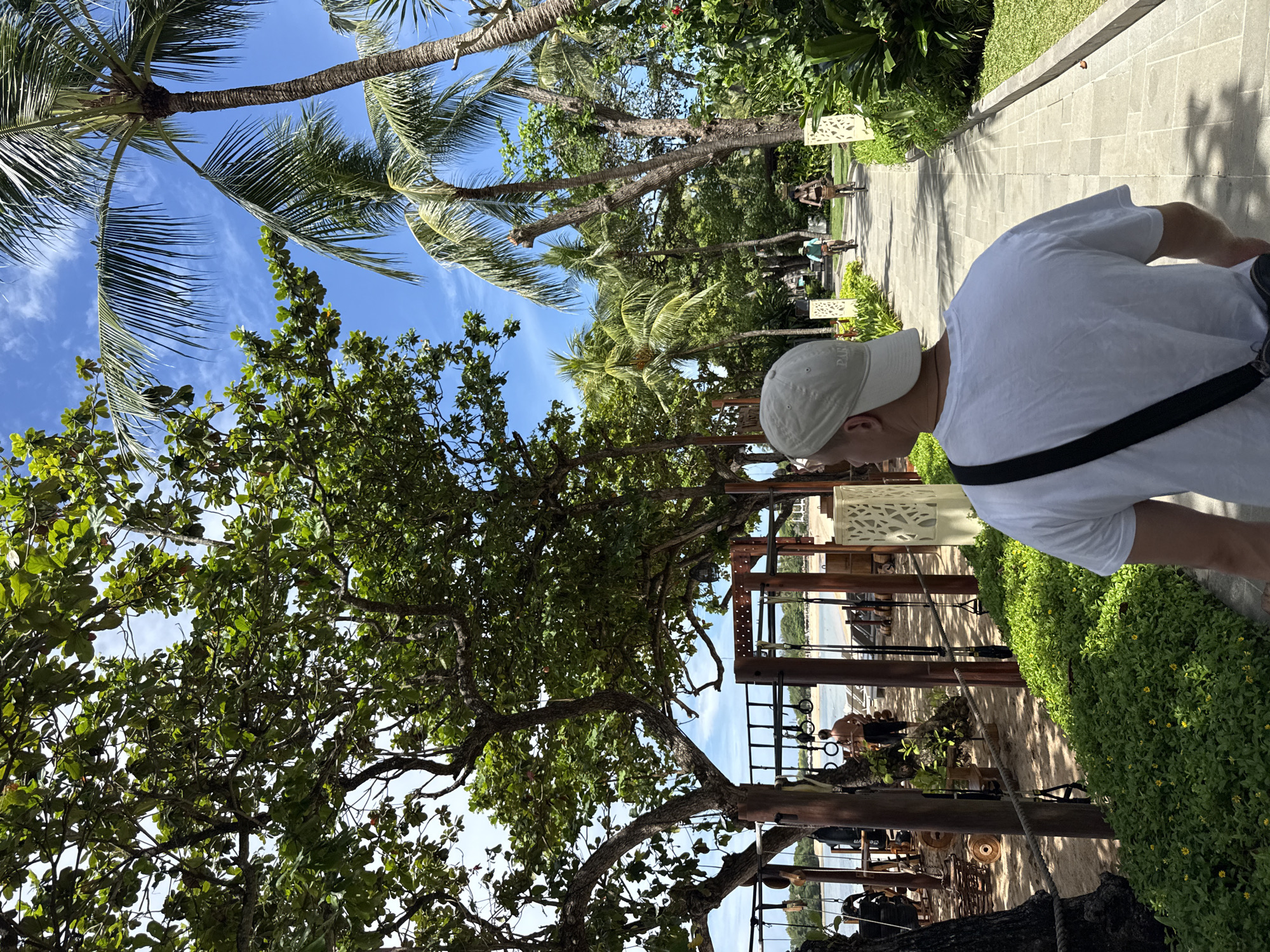 The image depicts a sunny day in Kuta Selatan, Indonesia. A person wearing a white T-shirt and a cap is walking on a stone pathway. Palm trees line the path, interspersed with other trees providing ample shade. In the background, people can be seen walking and cycling along the path. To the right, green bushes border the pathway. To the left, there is a view of a beach with wooden structures and people engaged in activities near the shoreline.