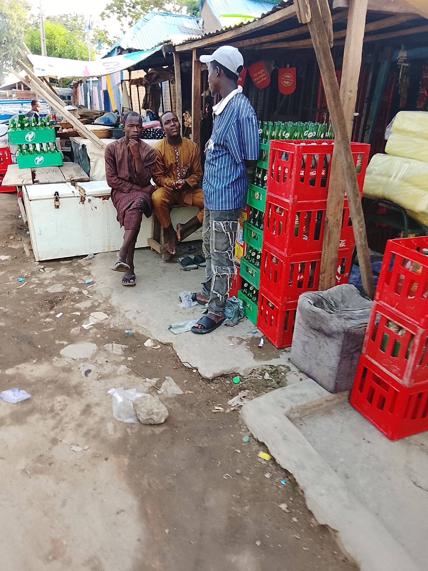 The image depicts an outdoor scene in Abuja, Nigeria, likely near a roadside market or informal business. Three men are visible: two are sitting on what appears to be a cooler or storage box, while the third is standing and talking to them. The seated men are wearing traditional-style clothing, one in brown and the other in yellow. The standing man is wearing a blue and white striped shirt, ripped jeans, and a white baseball cap.
Behind the standing man are stacks of red and green plastic crates filled with green glass bottles, likely soft drinks or beer. Yellow, wrapped items are stacked on top of one another. The setting includes a partially visible open-air structure with a corrugated metal roof, possibly a shop or stall. Debris and litter are scattered on the ground, indicating a somewhat unkempt environment.