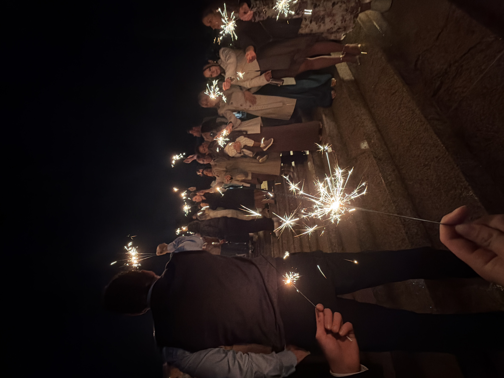 A nighttime outdoor scene depicts a group of approximately 15 individuals gathered on what appears to be a stone staircase or elevated platform. They are holding lit sparklers, which are emitting bright, glittering sparks. The people vary in height and dress, with some wearing jackets or coats. The background is dark, likely representing the night sky. Based on the architecture and the attire of the people, the location is Vila Real, Portugal. The individuals are interacting by standing close together and participating in the sparkler activity.