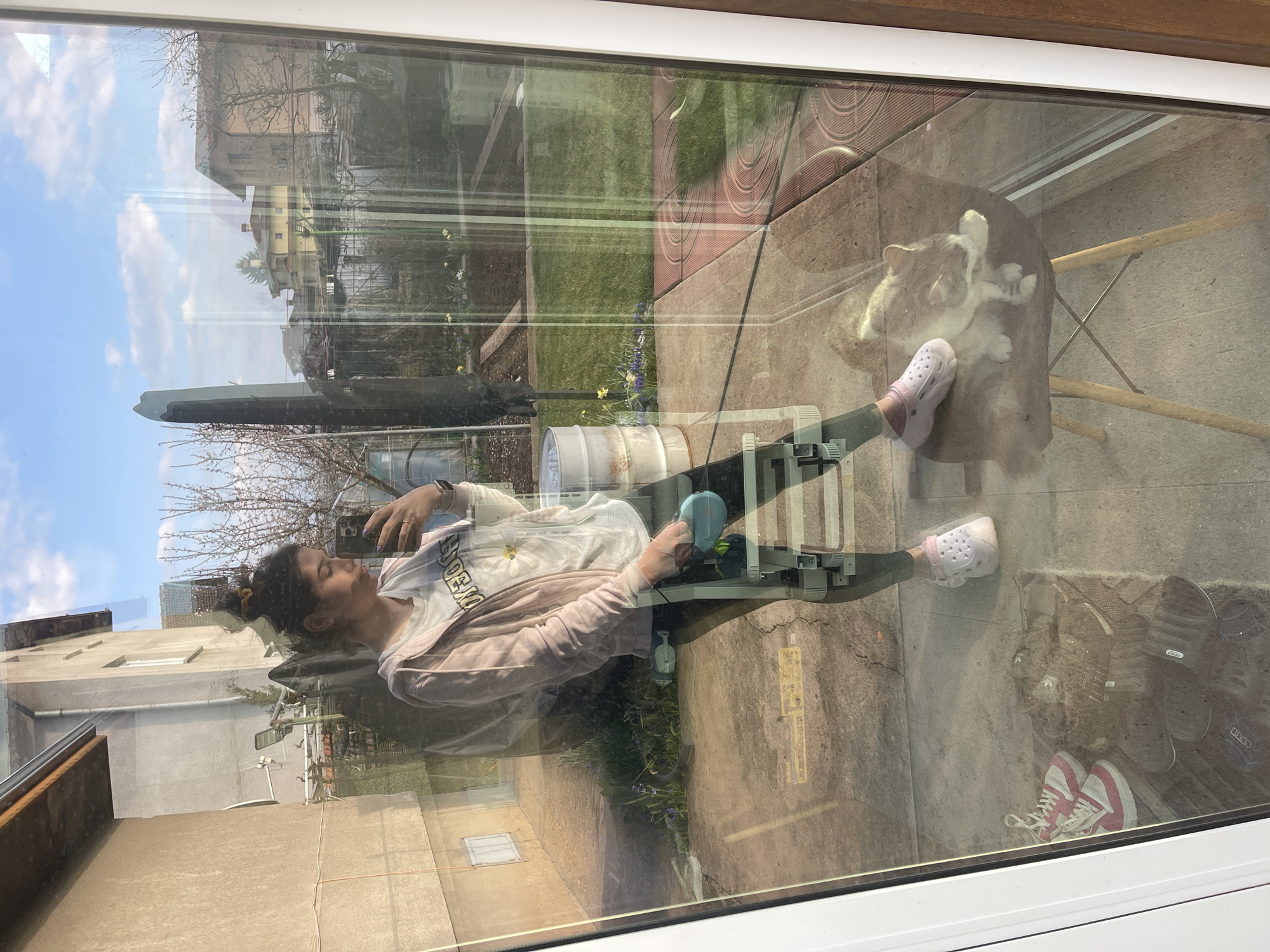 A woman is visible through a window, sitting outside in what appears to be a backyard or patio area. The reflections on the glass make it difficult to discern the exact location and details, but the background seems to show elements of an urban environment.
The woman is seated on what looks like a metal framed chair. She wears a light-colored sweatshirt, dark pants, and pink Crocs. She is holding a phone and appears to be taking a selfie. There is a cat or dog sitting on a chair nearby. There are other items scattered on the ground, including shoes and a barrel or container.
Given the presence of Cyrillic characters on a sign in the background, the image is likely taken in Bulgaria, specifically Sofia.