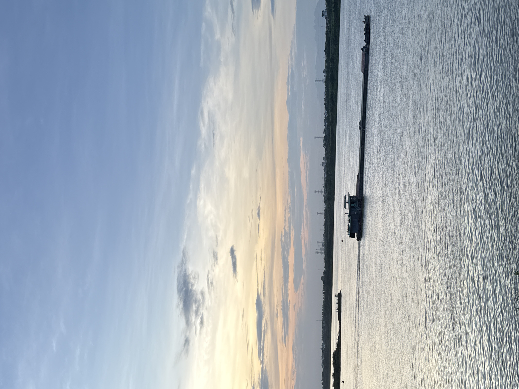 The image depicts a river scene in Hà Nội, Vietnam. A long barge, possibly transporting goods, is visible on the water. The background shows a shoreline with some vegetation and electricity pylons in the distance. The sky is partially cloudy with a sunset.
