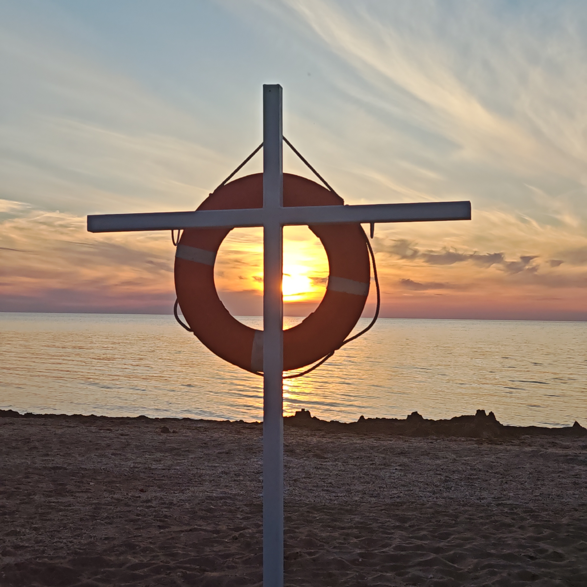 A forensic analysis of the provided image reveals a sunset scene captured from a sandy beach.
In the foreground, a prominent white cross-shaped structure is erected in dark, textured sand. The vertical post of this structure extends upwards from the bottom of the frame, intersecting with a horizontal bar positioned near its top. Suspended from the horizontal bar is an orange lifebuoy, which features two distinct white bands. The lifebuoy is precisely positioned such that the setting sun is centered within its circular opening.
The sun, a source of intense yellow-orange light, is partially obscured by the lifebuoy and the vertical post, creating a backlighting effect. The sky above transitions from a pale blue at the top to vibrant hues of light orange, yellow, and soft pink closer to the horizon. Wispy, elongated clouds are visible across the sky, particularly in the upper right and middle sections, illuminated by the sunset colors.
Below the horizon line, a calm body of water, likely a sea or ocean, stretches across the midground. The water's surface reflects the warm tones of the sunset, appearing golden and orange, with subtle ripples visible. The shoreline, separating the water from the sandy beach, is marked by an irregular, dark line composed of what appears to be small rocks or accumulated debris. The sandy beach in the immediate foreground is dark brown, indicating it is likely in shadow or wet.
There are no discernible human figures, animals, or other significant man-made objects present in the scene, which depicts a natural coastal environment at dusk. The location is in the city of Unknown, Unknown.
