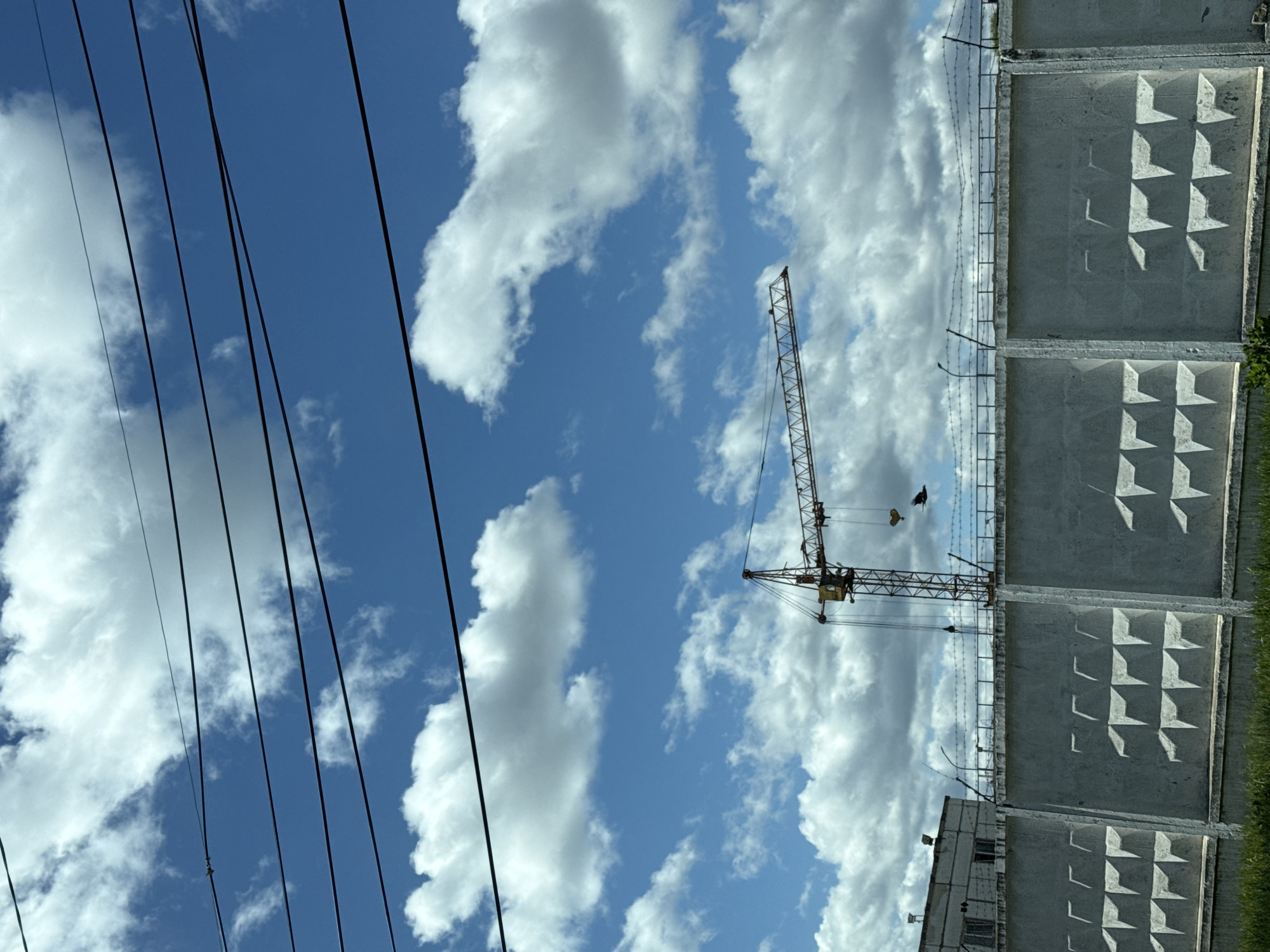 The image shows a view in Chaikovsky, Russia. Dominating the right side of the image is a tall concrete wall with a repeating pattern of recessed geometric shapes. Above the wall, barbed wire stretches horizontally. To the left of the wall, a construction crane stands against a backdrop of a blue sky dotted with fluffy white clouds. Power lines run diagonally across the left side of the image. A building is partially visible at the bottom of the image, adjacent to the concrete wall. The overall impression is one of a construction site next to an enclosed or restricted area, under a bright sky.