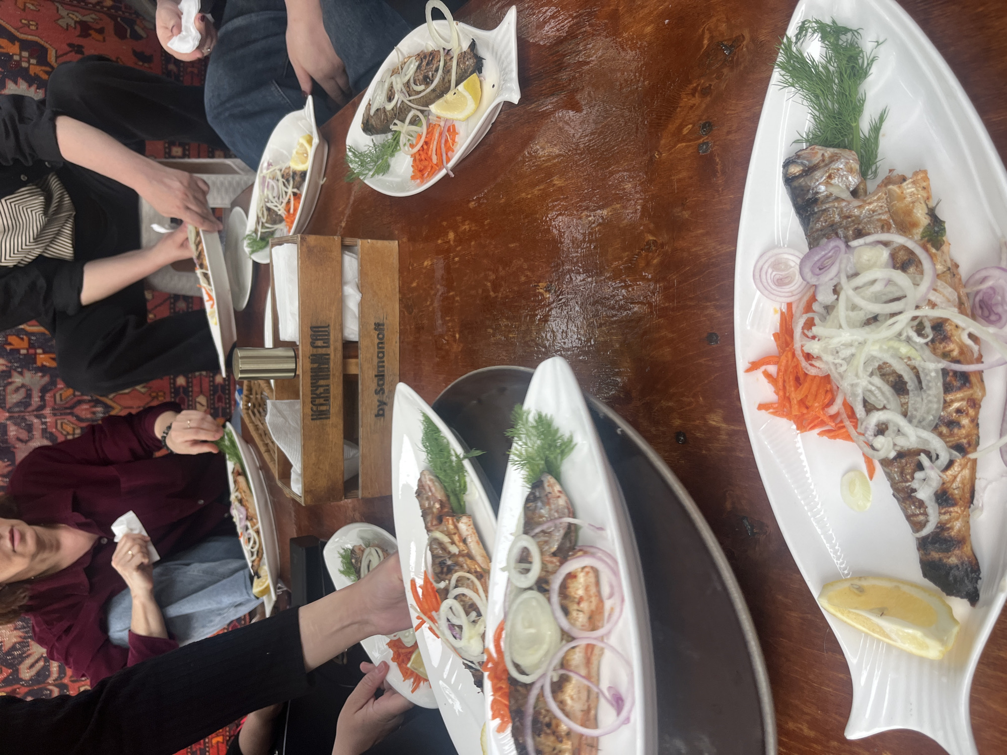 A tilted, medium shot depicts a gathering of people sharing a meal, potentially in Miatli, Russia. The focal point is the grilled fish served on white, fish-shaped platters, garnished with dill, red onion rings, grated carrots, and lemon wedges. Several plates are on a dark wooden table, with a wooden box labeled "by Salmanoff" in Cyrillic script. People are seated around the table, and hands can be seen reaching for the plates. A person on the left is holding a white napkin. The background includes a rug with an oriental pattern. The image suggests a casual dining experience, focusing on the presentation of the food.