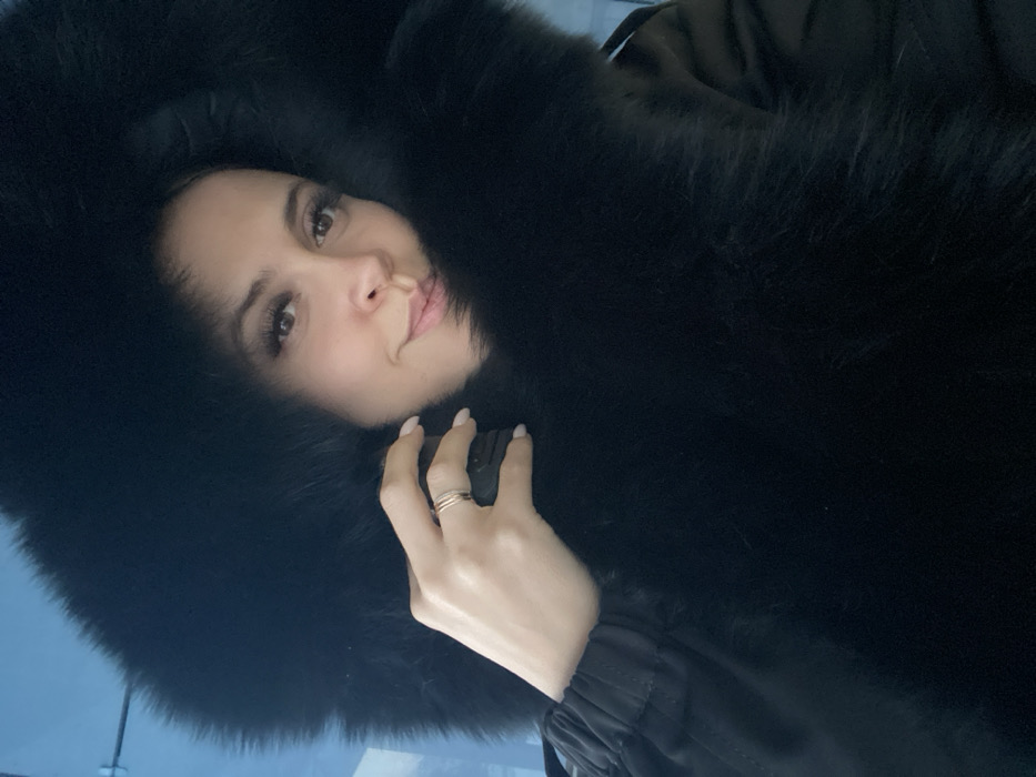 A female subject is depicted in a close-up, frontal portrait, likely a selfie. She has fair skin, dark hair, light brown eyes with prominent eyelashes, and light pink lips forming a subtle smile. Her face is framed by a voluminous, dense black fur hood of a dark, likely black, winter coat or parka. Her right hand, exhibiting light-colored fingernails and a three-band ring on the ring finger, is visible near her left shoulder, gripping a dark, indistinct object that appears to be part of her attire. The background is a blurred light blue, with a faint horizontal line visible in the upper left quadrant. The scene is located in the city of Chaikovsky, Russia.