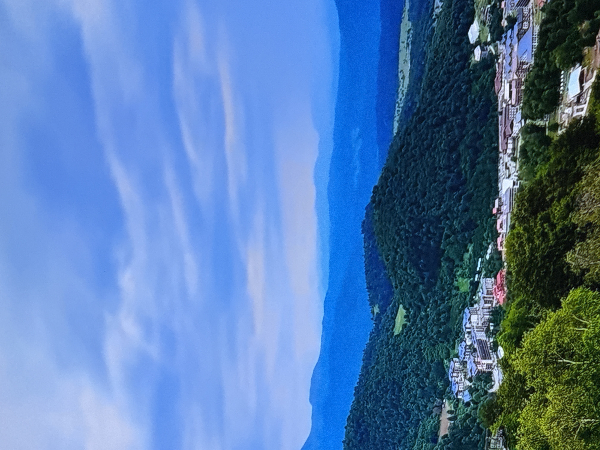 The image captures a panoramic view of a mountainous region with a town nestled within the valleys. The sky above is a vibrant blue, streaked with wispy white clouds, creating a sense of depth and openness. In the foreground, lush green trees dominate the scene, their leaves a rich emerald hue, suggesting a temperate climate and a thriving natural environment. Beyond the trees, the terrain slopes downwards, revealing a collection of buildings that form a small town or village. These structures are characterized by their varied roof colors, with some featuring terracotta tiles and others a deep blue, hinting at modern architecture interspersed with traditional elements. The town is surrounded by rolling hills covered in dense, dark green forests, their textures suggesting a rugged and untamed landscape. In the distance, layers of mountains fade into a hazy blue, creating a sense of vastness and solitude. The overall impression is one of natural beauty and tranquility, with human habitation harmoniously integrated into the mountainous terrain.