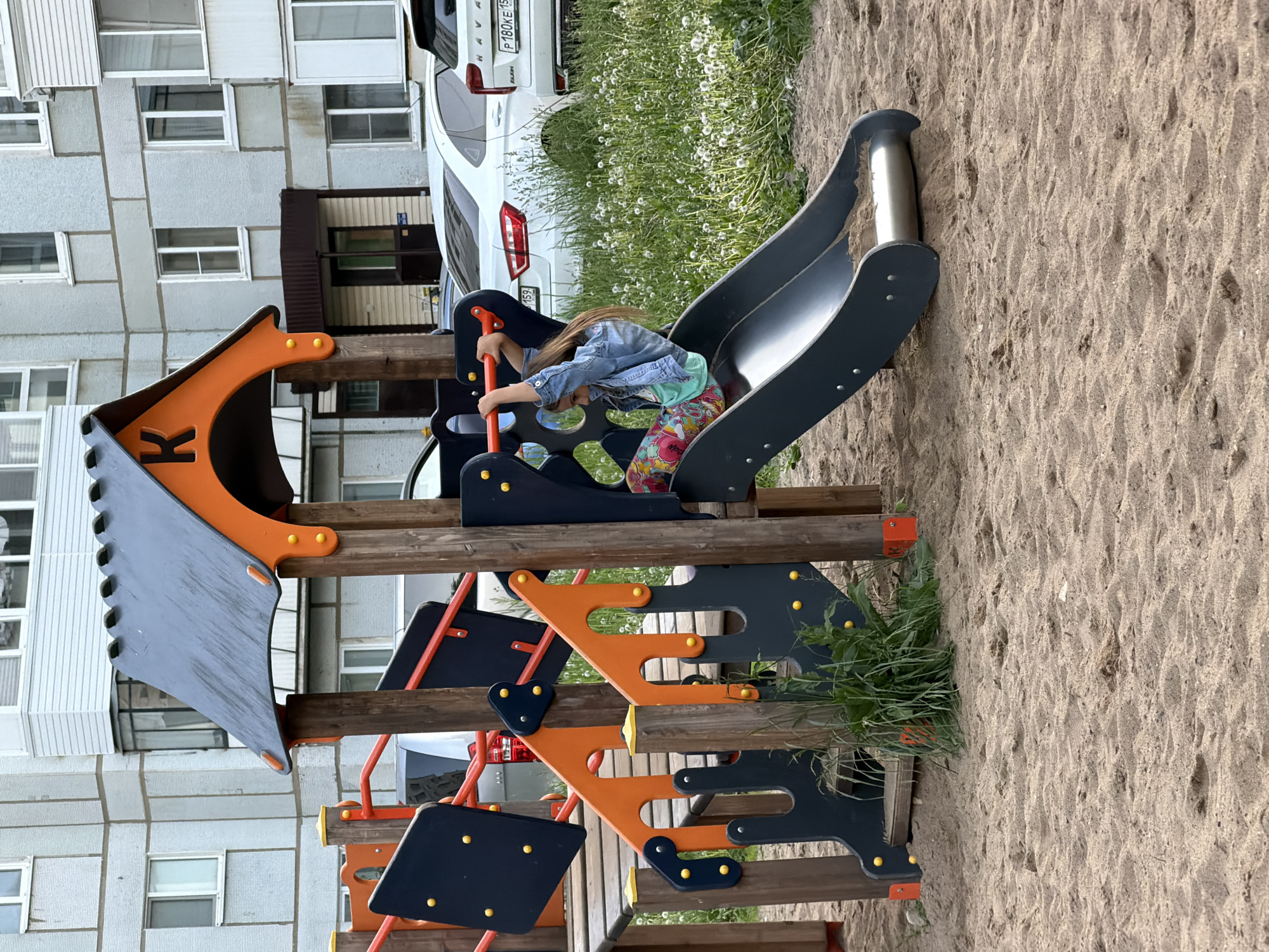 The image depicts a playground scene in Chaikovsky, Russia, during what appears to be daytime under diffused lighting. A young child with light brown hair, wearing a denim jacket, a light green top, and colorful patterned leggings, is situated on a dark gray slide of a playground structure. The child is holding onto an orange handrail, appearing focused as they either prepare to slide down or climb onto the slide.
The playground structure features dark gray and orange elements, including various climbing components and a slide, supported by sturdy wooden beams. The ground beneath and around the playground is sandy, showing ripples and footprints.
In the background, a large, multi-story apartment building with numerous rectangular windows is visible on the left, characteristic of an urban residential area. To the right and behind the playground, a white SUV is parked, identified as a Haval by text on its trunk, with the visible Russian license plate "P180KE 159." A strip of green grass populated with many white dandelion puffs separates the parked car from the play area.