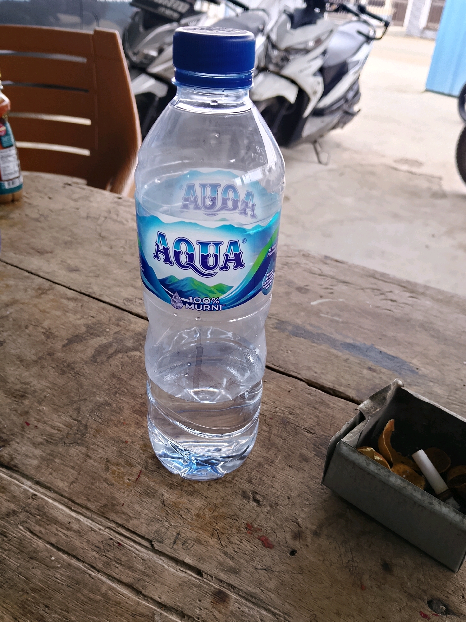 A clear plastic bottle of Aqua brand mineral water with a blue cap and a label featuring a mountain and water design is positioned on a rustic wooden table. The bottle is about two-thirds full of water. To the right of the bottle, an ashtray made of metal contains cigarette butts and some orange peels. In the background, out of focus, a motorcycle is visible, suggesting an outdoor or open-air setting. The wooden table appears worn and weathered.