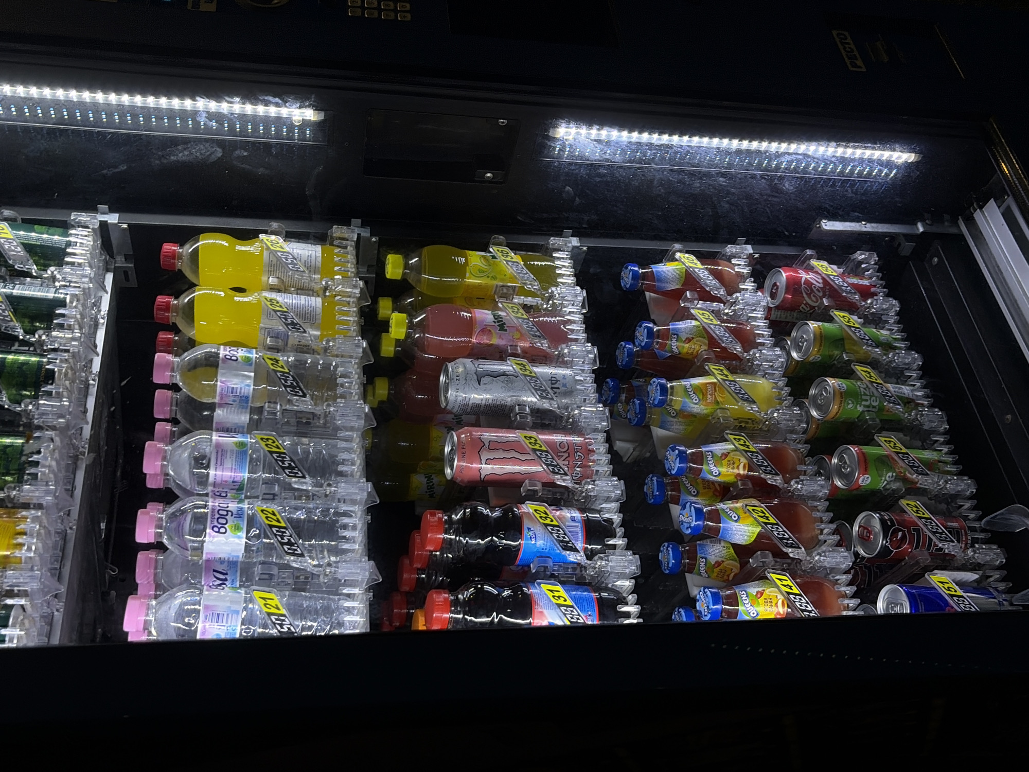 An upright, interior shot of a brightly lit, multi-shelf vending machine displays a variety of beverages. The machine is filled with numerous bottles and cans of drinks, neatly organized on transparent plastic racks, each with a yellow price tag indicating a numerical code. The top shelf holds clear bottles with pink caps, followed by yellow-capped bottles. The middle shelves feature dark-colored bottles, a large red Monster Energy can, and other cans with colorful labels. The bottom shelves showcase more cans and smaller bottles. The machine's interior is illuminated by bright white LED lights lining the right side, casting reflections on the glass and products. A dark, textured surface forms the right side of the machine, featuring a keypad and a slot, possibly for payment or dispensing. The overall environment appears to be indoors, possibly in a public space like a hotel, restaurant, or shop in the city of Obzor, Bulgaria. There are no people or animals visible in the image.