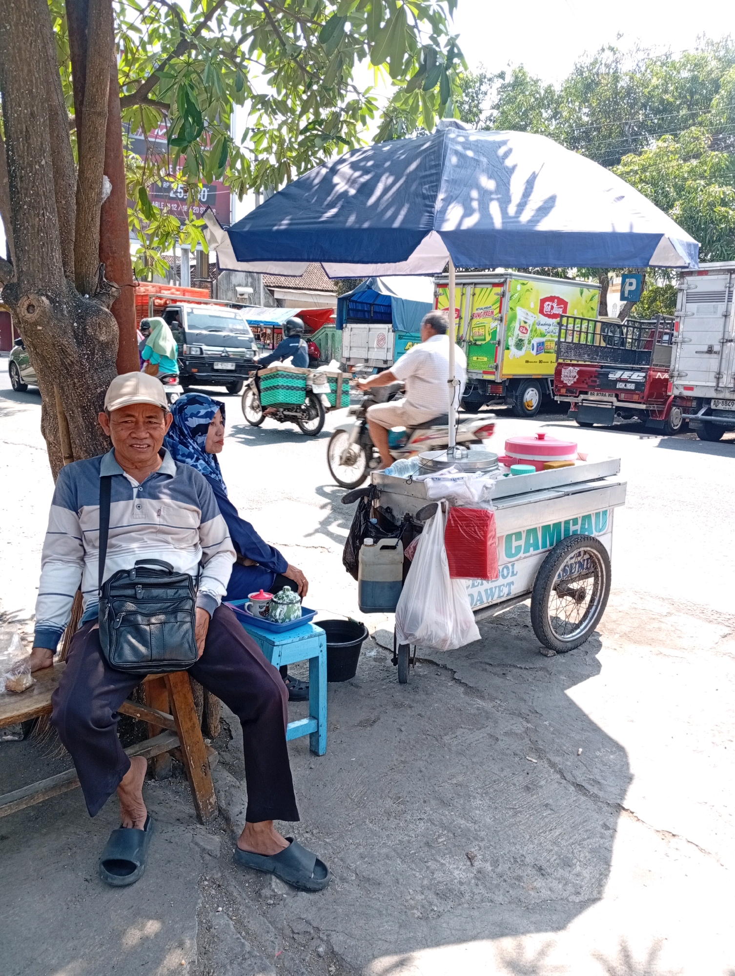 In Playen, Indonesia, a man and a woman are sitting on a bench under a tree on a sunny day. The man is wearing a striped shirt and a cap, with a black bag slung over his shoulder. The woman is wearing a blue hijab and a dark blue top. They are positioned in front of a food cart, which has the words "CAMCAU" and "EMPOL DAWE" written on it. The cart is equipped with a pink lid and a red container, and it has two wheels. In the background, there are vehicles, including trucks and motorcycles, and trees lining the street. The scene appears to be a typical street view in a bustling Indonesian town.