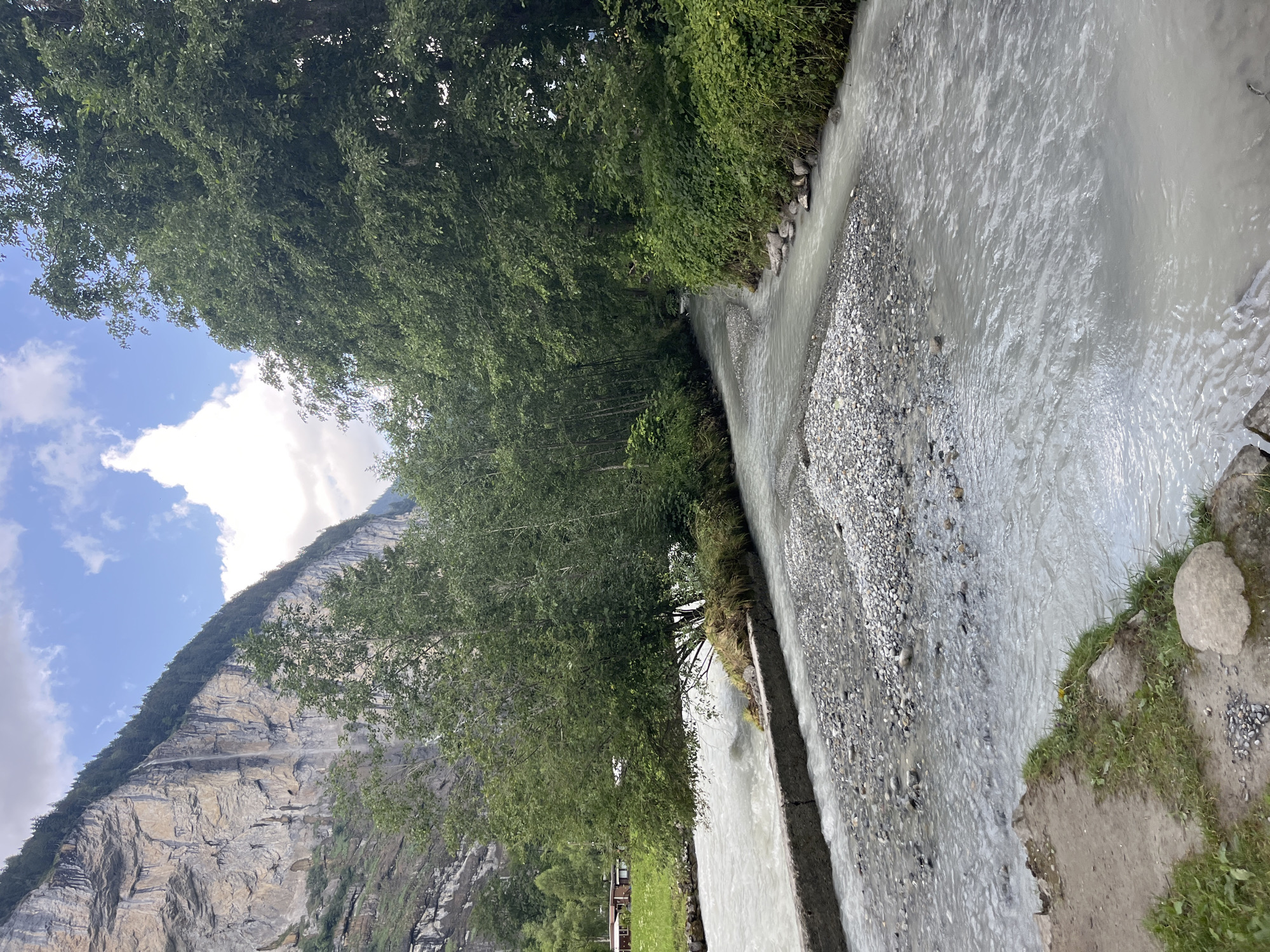 The image depicts a scenic view in Wengen, Switzerland. A wide, milky-white river flows through a valley, bordered by lush green trees and a steep, rocky cliff face. The riverbed is visible in places, with numerous grey and white pebbles. A concrete structure, possibly a weir or a retaining wall, runs along the left bank of the river in the foreground. On the far left bank, a small patch of grass and gravel is visible, with a single large, light-colored rock. In the middle ground, trees line both sides of the river, with a dense forest on the right. The cliff face, covered in scattered trees and vegetation, dominates the background on the left. A thin waterfall cascades down the cliff face. The sky is partly cloudy with patches of blue. There are no people or animals visible in the image.