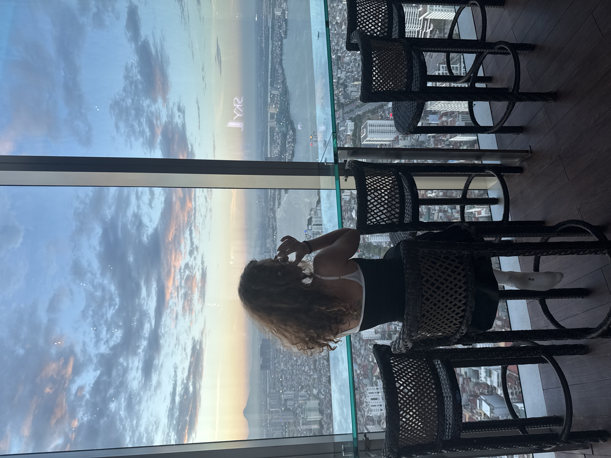 A woman with curly brown hair is seated at a bar overlooking a city at dusk. She is wearing a black and white tank top and white socks. The bar has a glass top and is lined with four dark woven chairs. In the foreground, the chairs and wooden floor are visible. The background shows a sprawling city with many buildings and a large body of water. Distant mountains are visible on the horizon. The sky is filled with clouds and painted with hues of orange, pink, and blue, indicative of a sunset or sunrise. A reflection of a logo that appears to read "SKY" is visible on the window. This scene is likely from a high-rise establishment in Hanoi, Vietnam, given the architectural style and the reflection of the "SKY" logo, which could correspond to a Sky Bar.