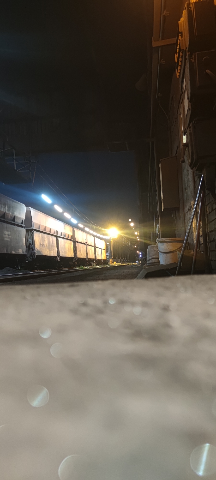 A train of freight cars is parked on railroad tracks at night. The train is illuminated by rows of lights along its side and a bright yellow street lamp further down the track. On the right side of the image, a brick wall is visible with electrical boxes and conduits attached. Two buckets are placed on a surface near the wall. The foreground is blurred with out-of-focus circular bokeh lights, suggesting a low-angle shot possibly from a wet or reflective surface. This scene appears to be in Salzgitter, Germany, likely within an industrial area.