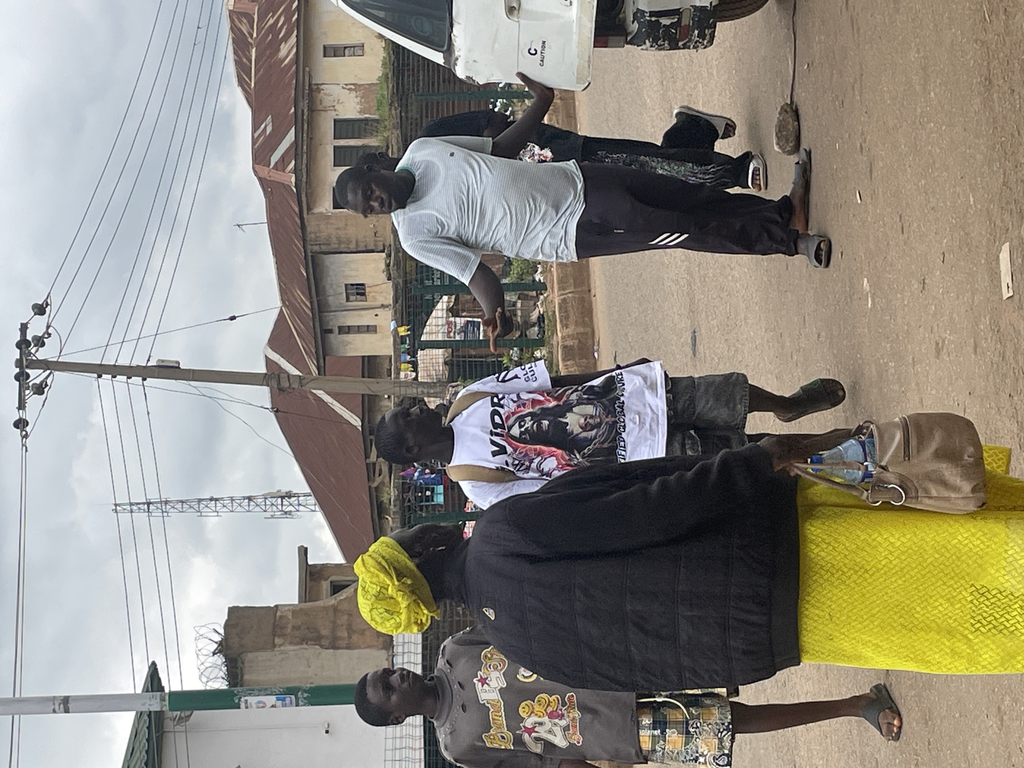 In Bukuru, Nigeria, a group of people are gathered on a street. A man in a light blue t-shirt and dark pants gestures outwards while facing away from the camera. To his left, a person wearing a black jacket and a bright yellow headwrap stands facing the man, holding a tan purse with a water bottle inside. To the left of these two, a young man in a gray t-shirt with yellow smiley faces printed on it stands looking to his left. Further to the left, another young man in a white t-shirt with a graphic print is partially visible. A white vehicle is parked on the right side of the frame. The street is paved, and buildings with corrugated metal roofs line the background, along with utility poles and wires. A cell tower is visible in the distance.