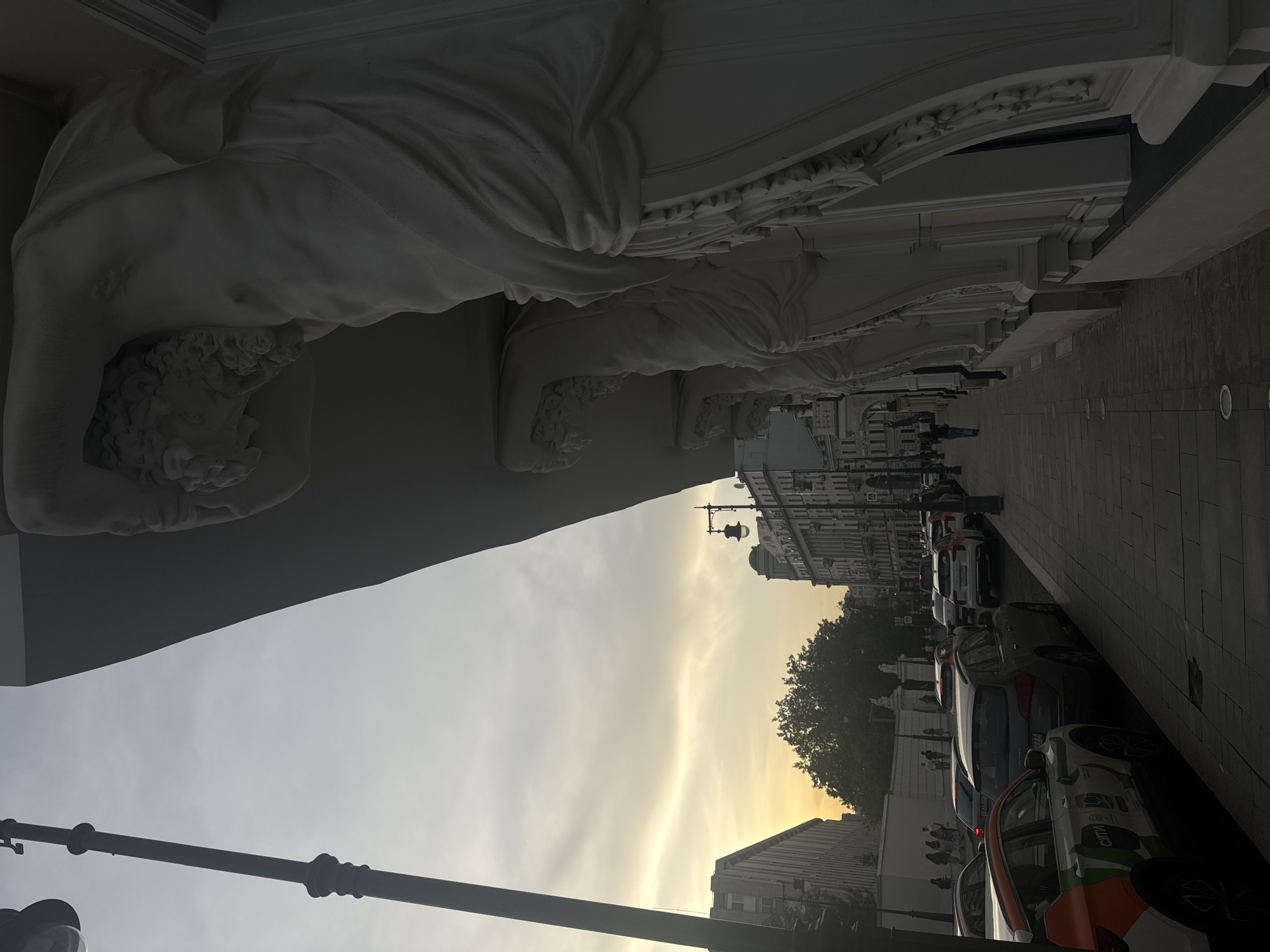 The image shows a street in Moscow, Russia. Along the top of the image, there is a decorative architectural feature with a white statue resembling a man. The streetscape shows buildings on either side, a row of parked cars, and a sidewalk. People are walking on the sidewalk. The sky appears hazy with a gradient of light from a setting sun. A lamppost is visible on the left.
