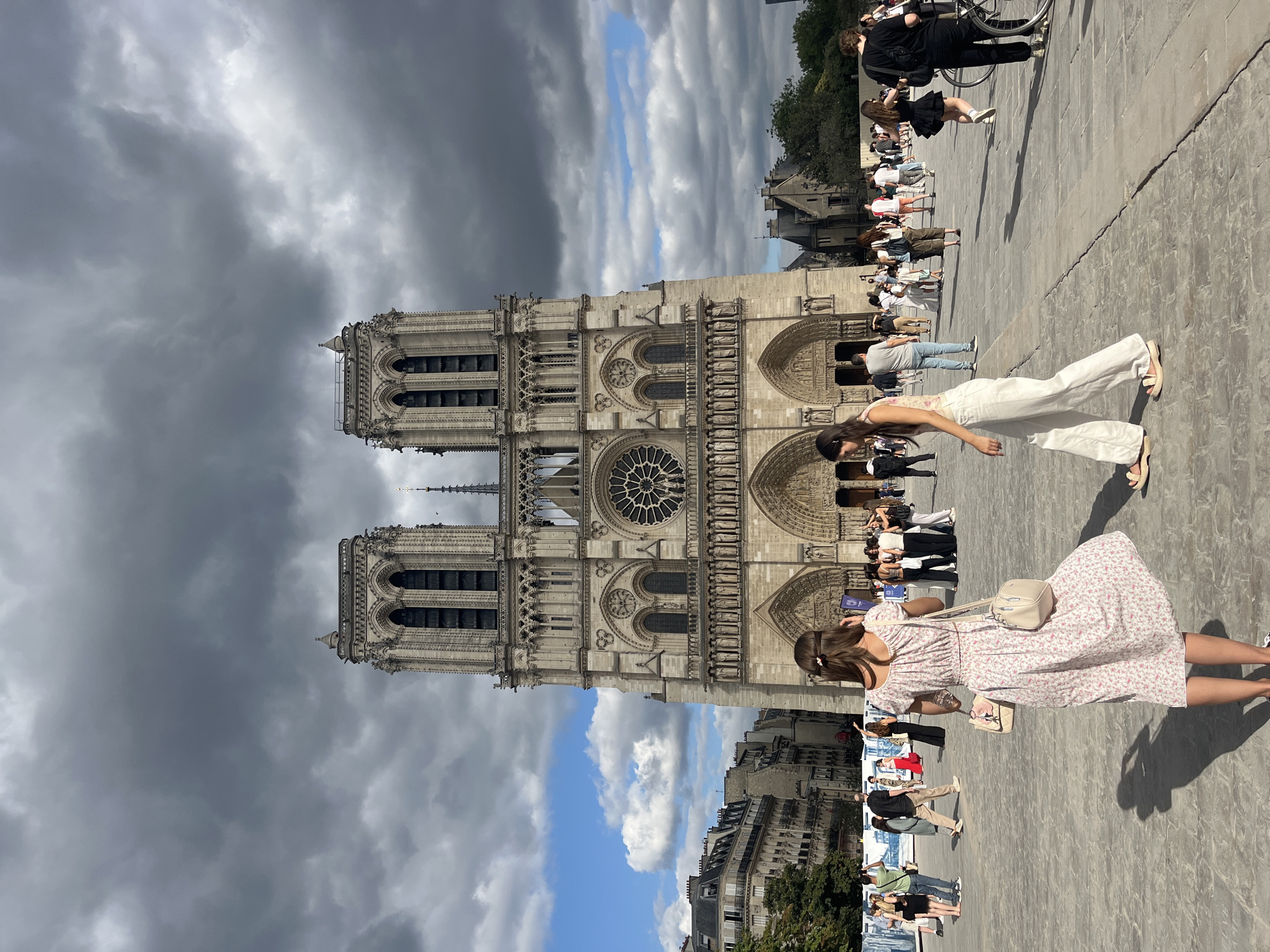 This is a full-body, eye-level shot of Notre Dame Cathedral in Paris, France, under a dramatic, cloudy sky. The facade of the cathedral is the central focus, with its two large towers, rose window, and multiple arched entrances. A large plaza in front of the cathedral is populated with numerous people. In the foreground, a young woman in a white floral dress is captured from behind, facing the cathedral and holding two bags. To her right, another young woman walks away from the camera, wearing white linen pants and a tank top. Other individuals are seen scattered across the plaza, some walking, some standing in groups, and one person on a bicycle on the right side of the frame. The ground is paved with cobblestones, and buildings line the background to the left of the cathedral. The overall scene depicts a sunny day with a mix of sunlight and heavy cloud cover.