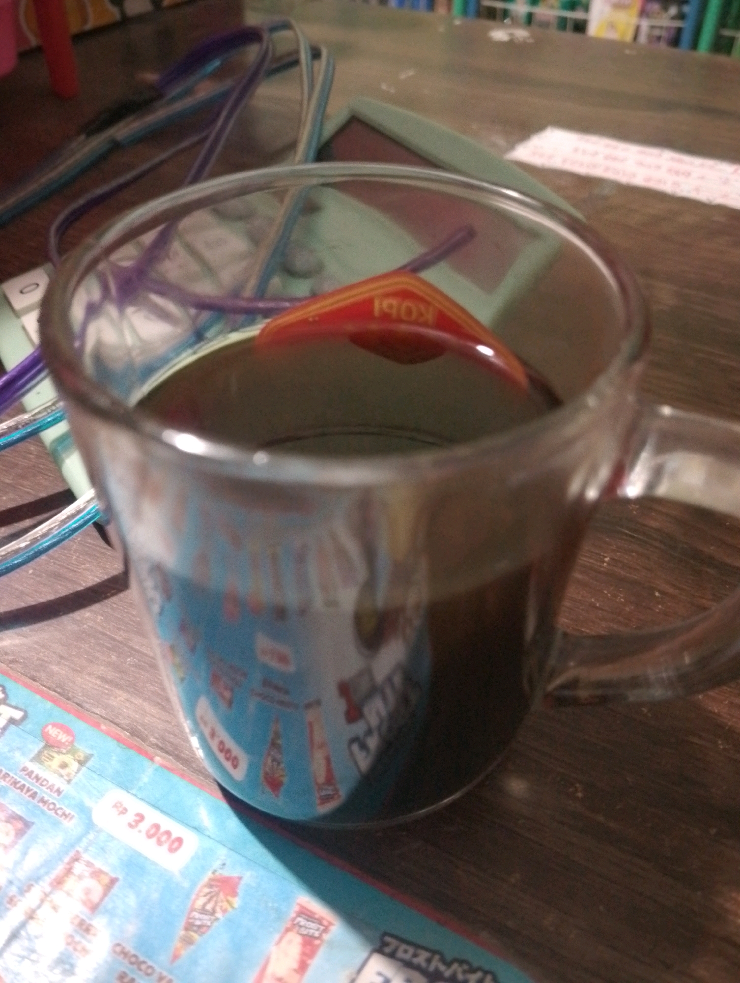 A clear glass mug filled with dark liquid, likely coffee, sits on a wooden surface. The mug is partially filled, with the liquid reflecting light. A red and yellow object, possibly a coaster or a label, with the text "KOPI" visible in reverse, is submerged in the liquid. Various colored wires, purple and blue, are intertwined around the mug and extend out of the frame. In the background, a light green object, perhaps a book or a folder, is visible. Below the mug, a portion of a colorful item, possibly packaging or a magazine, is visible. This item features text in various fonts and colors, including the text "NEW!" and "Rp 3.000". The lighting suggests an indoor setting, possibly a cafe or a home environment. The location is not specified in the image.