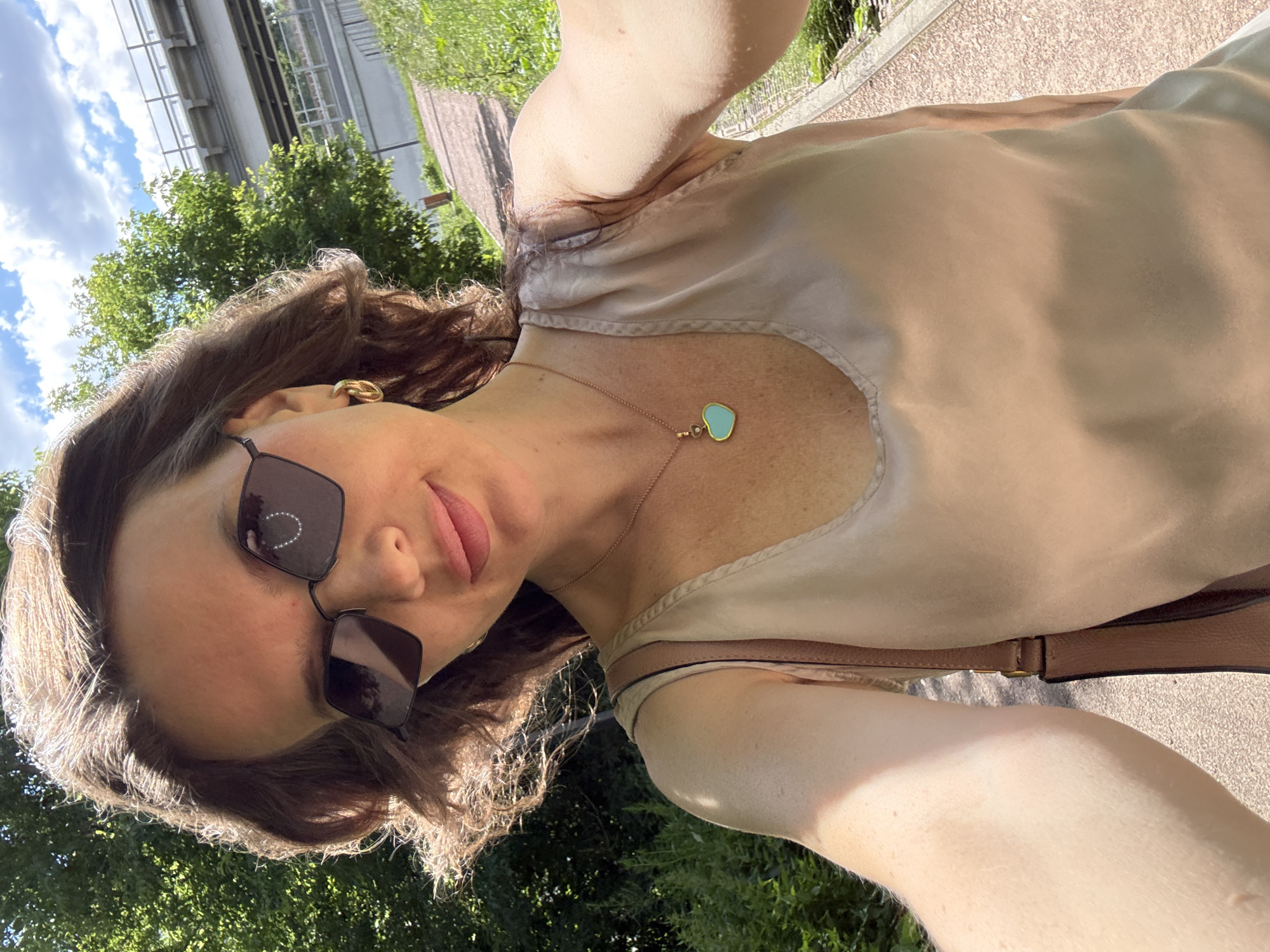A woman is taking a selfie outdoors, outdoors in a park-like setting. She is wearing sunglasses, a light-colored top, and a necklace with a heart-shaped pendant. Her hair is brown and wavy. Behind her, there are trees and a blue sky with clouds. The ground is a paved path. The scene appears to be during the day, with bright sunlight. There is no visible text in the image.