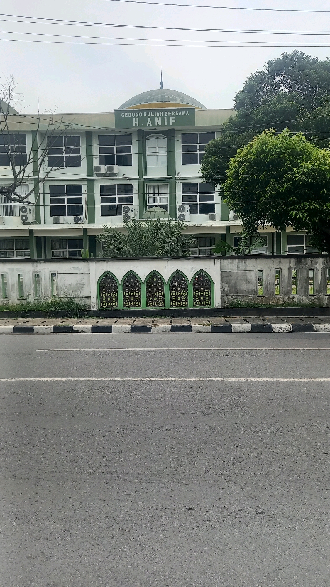 The image captures the facade of the "GEDUNG KULIAH BERSAMA H. ANIF" building, likely a university lecture hall, located in the city of Medan, Indonesia. The building is light green with white trim and features multiple floors with numerous windows and air conditioning units visible. A domed structure, characteristic of Islamic architecture, is positioned centrally atop the building, topped with a spire. In front of the building, a white wall with ornate green archways and intricate metalwork serves as a decorative barrier. Beyond the wall, a paved road with a distinct black and white striped curb runs horizontally across the foreground, indicating it is a street in a developed area. Tall trees, some with lush green foliage and others bare-branched, frame the sides of the building. The sky above is overcast, suggesting a cloudy day. The scene is devoid of people or any discernible activity.