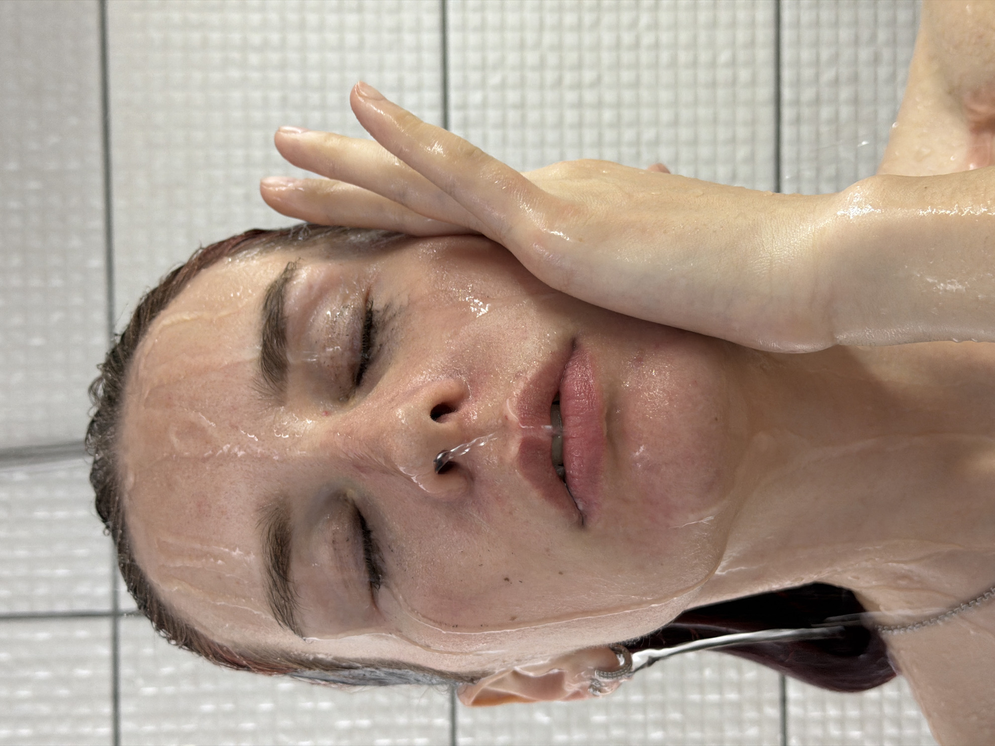 A fair-skinned person with dark hair is depicted in a close-up image, their face wet as if from a shower. Their eyes are closed. The person's right hand is raised, with the fingers slightly spread and touching their forehead. Water appears to be running down their face. A small piece of jewelry is visible on the ear. A necklace is seen as well. The background appears to be tiled. There is no information in the image indicating the location of Chaikovsky, Russia.