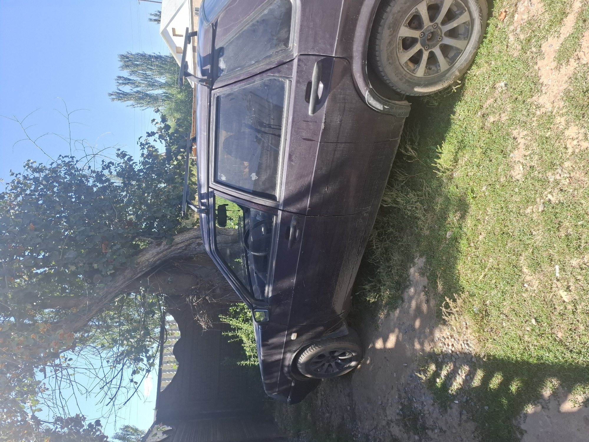 The image shows a dark-colored car parked on a patch of grass and dirt next to a wooden fence and a large tree. The car appears to be a hatchback with a roof rack. The environment suggests a rural or semi-rural setting with greenery and a clear blue sky. The lighting indicates it is daytime, likely mid-morning or afternoon, with shadows cast by the tree and fence. The car is dusty, suggesting it has been used recently or exposed to the elements. In the background, there are more trees and the roof of a building. The overall scene is static, with no visible people or activity. This scene is located in the city of Jeti-Ögüz, Kyrgyzstan.