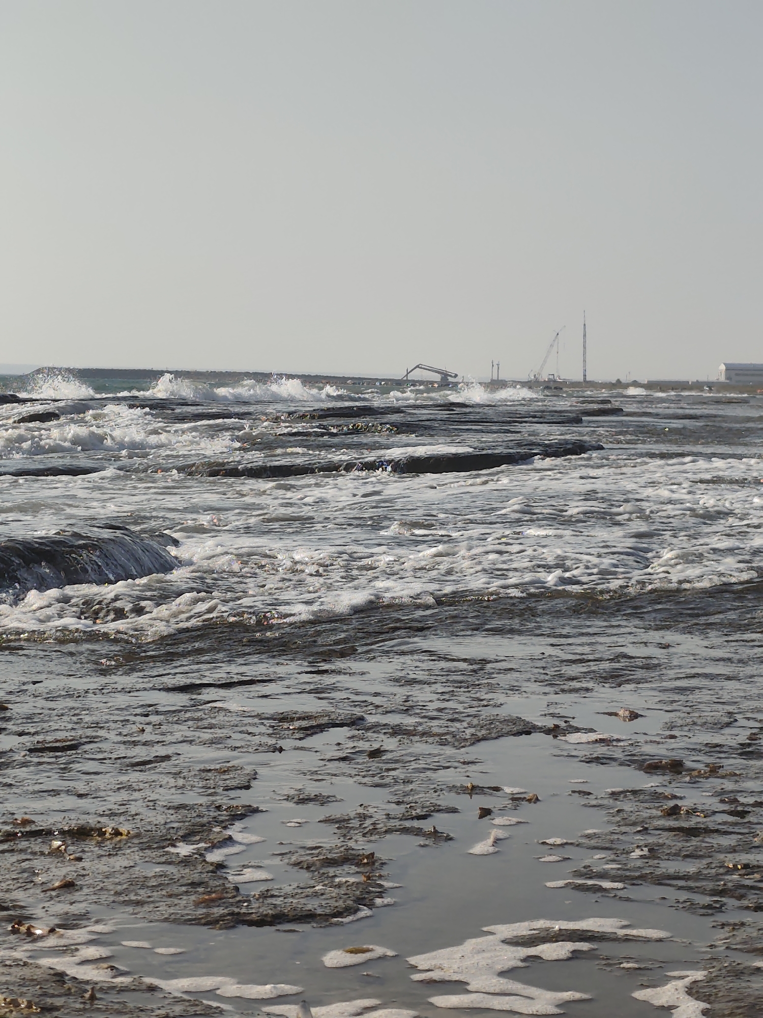 The image depicts a rocky shoreline with waves crashing onto the rocks. In the background, industrial structures are visible, including cranes and a tall tower, suggesting a port or industrial area. The sky is clear and overcast. There are no people or animals visible in the image. The foreground shows a muddy and rocky terrain with some foam from the waves. The scene appears to be located near the city of Saiyn Şapağatov, Kazakhstan, which is known for its industrial port on the Caspian Sea.