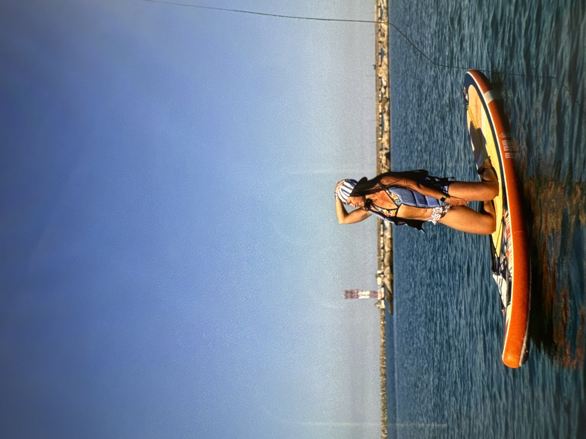 A woman in a bikini and a striped hat is sitting on a paddleboard on the water. She is wearing a life vest over her bikini. Her right arm is raised and she is touching her hat with her hand. The paddleboard is orange, yellow, and white with some colorful designs on it. The water is blue and appears calm with some ripples. In the background, there is a shoreline with buildings and a tall, red and white striped tower, likely a lighthouse or marker. The sky is a clear, light blue, suggesting a bright and sunny day. The scene appears to be a recreational activity on a clear day, likely during daylight hours. The woman's posture might suggest contemplation or a moment of rest. The text on the paddleboard is partially visible and appears to include "JBL SOUND SYSTEM" and possibly "ALMA."